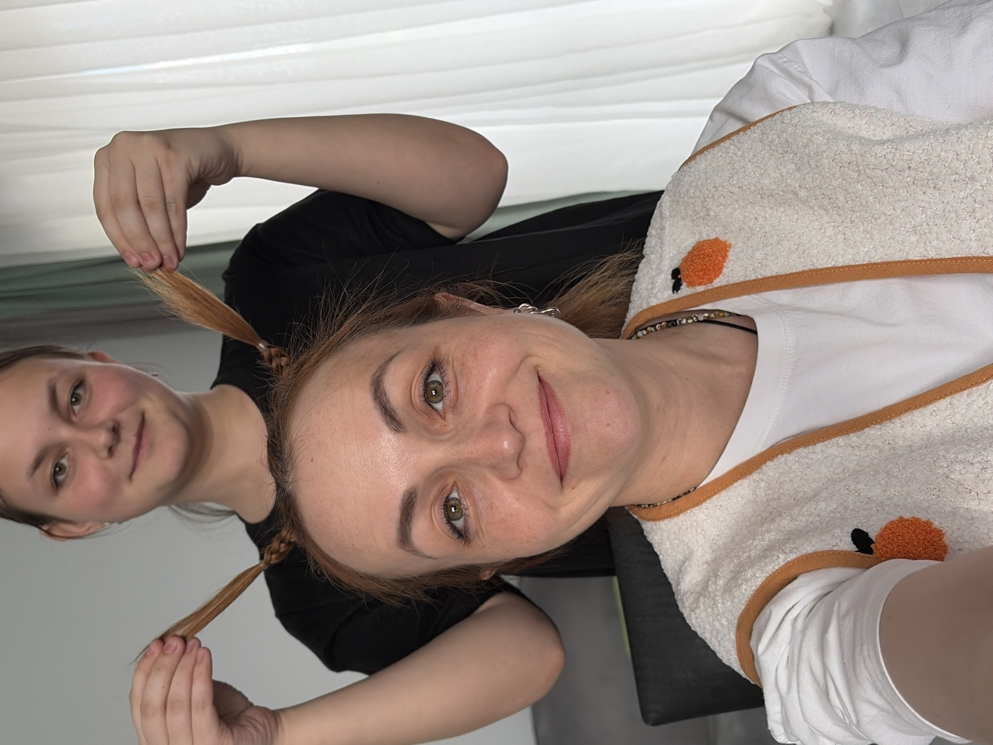 A woman with light skin and reddish hair is smiling while facing the camera. She has braided pigtails. A second person with light skin and dark hair is braiding the woman's pigtails. The woman is wearing a white shirt, and a white and orange cloth is draped around her. The second person is wearing a black shirt. The setting appears to be indoors. The city of Ольховка, Russia cannot be confirmed from this image.