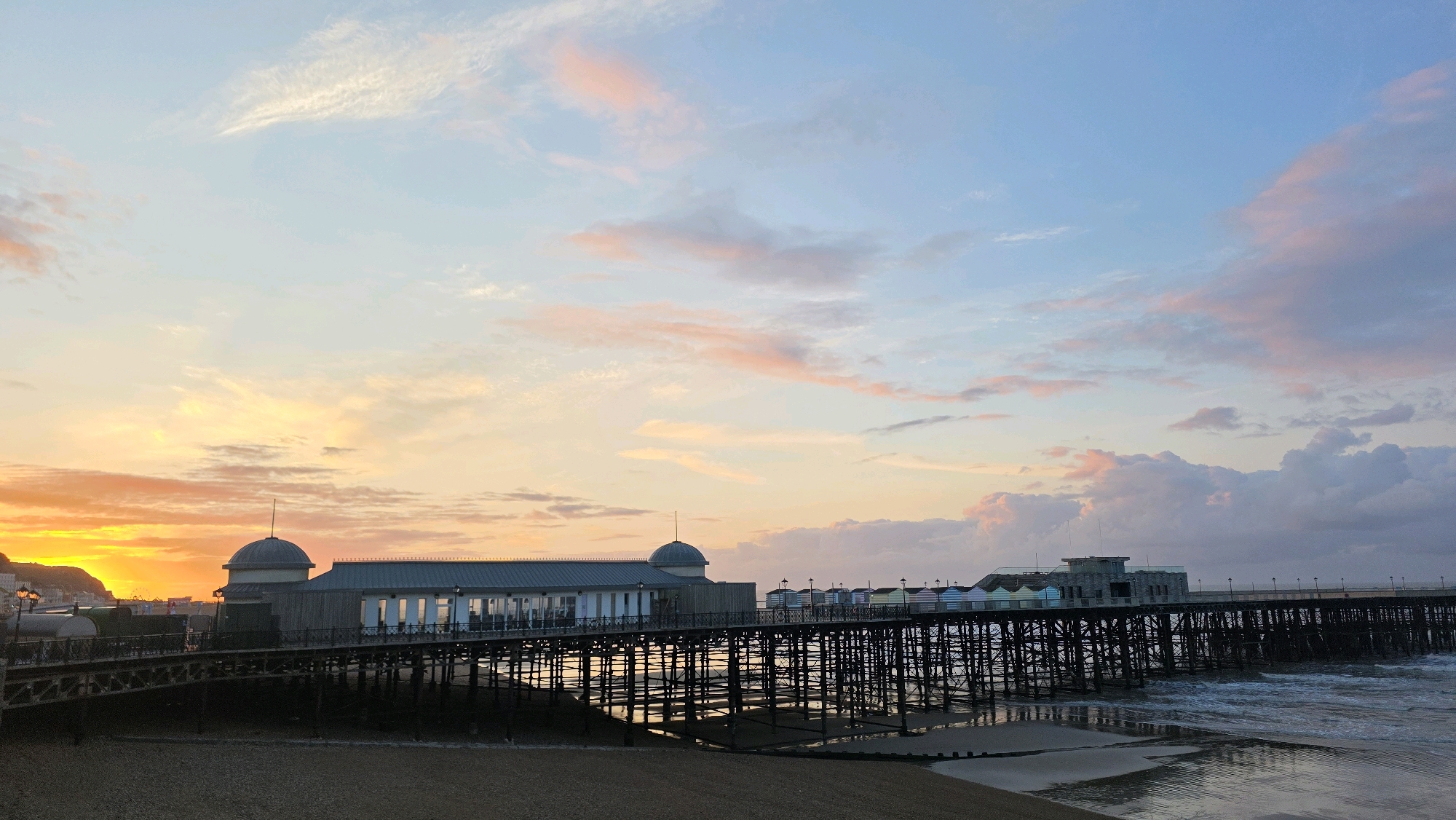 The image depicts the pier in St. Leonards-on-Sea, United Kingdom, at sunset. The sky is a gradient of blue and orange, with wispy clouds catching the fading sunlight. The pier extends out over the ocean, supported by numerous wooden pilings. At the shore end of the pier, there is a long, white building with a domed roof. Further along the pier, there are a row of colorful beach huts. The sea is calm with gentle waves lapping at the shore. The beach itself is composed of pebbles and sand. The overall scene is tranquil and picturesque. There are no people visible in the image.