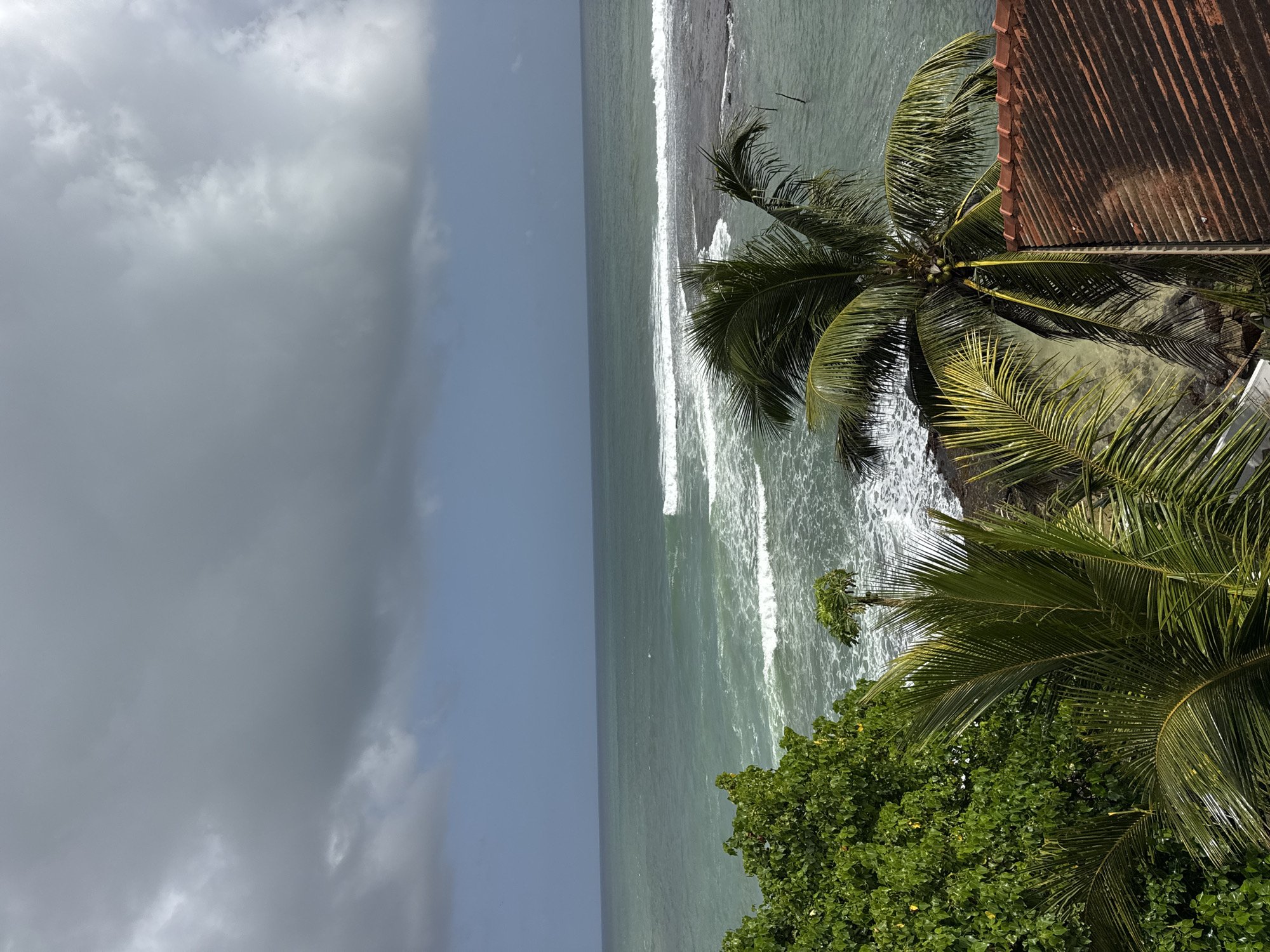 The image shows a view of the ocean from an elevated position in Goviyapana, Sri Lanka. The sky is overcast with dark, dramatic clouds, suggesting impending rain. The sea is a deep turquoise color, with white waves breaking against a rocky shore and a submerged reef. In the foreground, lush green trees and palm fronds frame the scene, with a tiled roof of a building visible on the right. The overall impression is one of a tropical coastline under a stormy sky. There are no people or animals visible in the image.