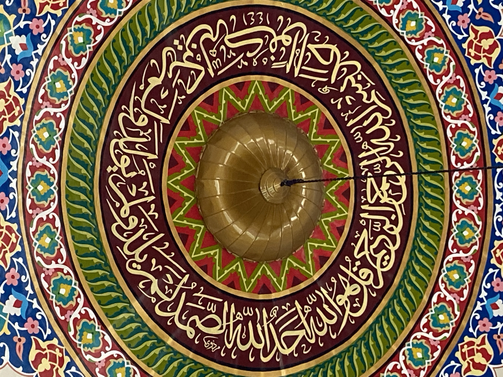 This image depicts the interior ceiling of a building, likely a mosque, featuring intricate Islamic calligraphy and geometric patterns. The calligraphy, rendered in gold, is circular and concentric with the dome at the center. Surrounding the calligraphy are concentric bands of color: deep red, green with a wave-like pattern, and an outer band with floral motifs in blue, red, yellow, and white. A golden, ribbed dome is visible in the upper center of the image, with a black cord hanging from it. The overall impression is one of rich decoration and religious significance. There are no people visible in the image. This ceiling is located in the Masjid Raya Islamic Center in Lhokseumawe, Indonesia.