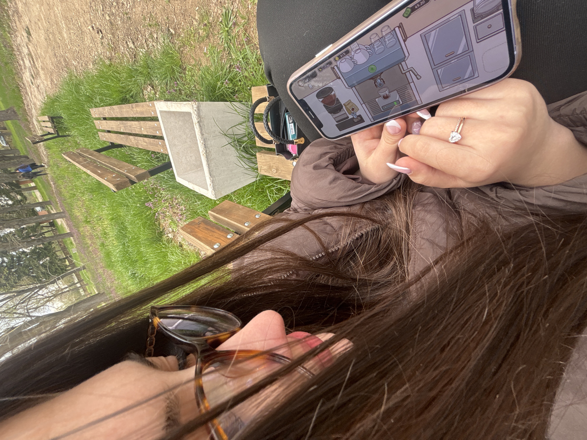 The image, taken from an elevated perspective, shows a person seated on a wooden park bench with a concrete planter nearby. The bench is located in a grassy area within a park, where other benches and trees are visible in the background. The location might be Lehchevo, Bulgaria, judging from the environment and landscape.
The person's torso, brown jacket, and dark hair are prominent in the foreground. A pair of tortoiseshell-framed glasses rests near the person's face. The person is holding a smartphone displaying a cartoon image of a coffee-making setup. A hand with manicured nails and a diamond ring is also visible, overlapping the phone screen. A small black purse sits on the bench next to the person.