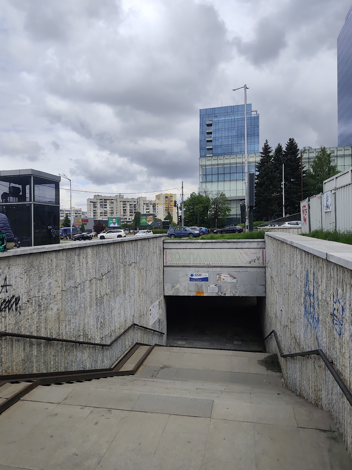 The image depicts an entrance to an underpass. The stairs are made of stone and have metal handrails. The walls of the underpass are made of a rough, light-colored material, possibly concrete or stone, with graffiti. Above the underpass is a road with several cars parked or driving along it. In the background are several buildings, including a modern, high-rise building with glass windows and a block of flats. The sky is cloudy. A sign with "GSM" written on it can be seen in the underpass entrance.