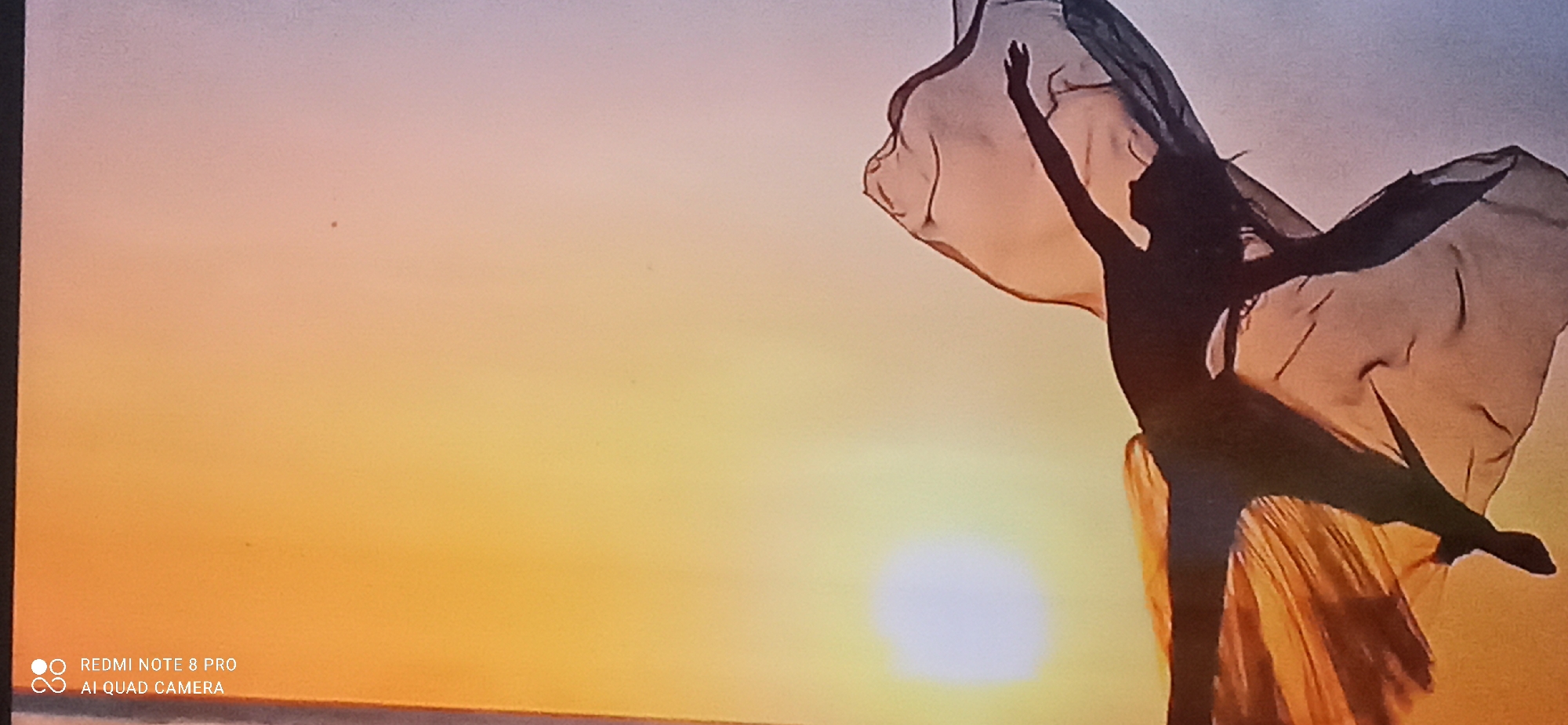 The image shows a silhouette of a person, likely a dancer, against a sunset sky. The dancer is in mid-pose, with one arm extended upwards and the other bent at the elbow. They are wearing flowing garments that are translucent and catch the light. The background is a gradient of colors, transitioning from a soft pink at the top to a warm orange and yellow towards the horizon, indicating a sunset. The sun itself is visible as a bright, diffused orb. In the lower left corner, there is a watermark with the text "REDMI NOTE 8 PRO AI QUAD CAMERA". The image is taken from a low angle, emphasizing the vastness of the sky and the dancer's movement. The overall impression is one of grace and freedom.