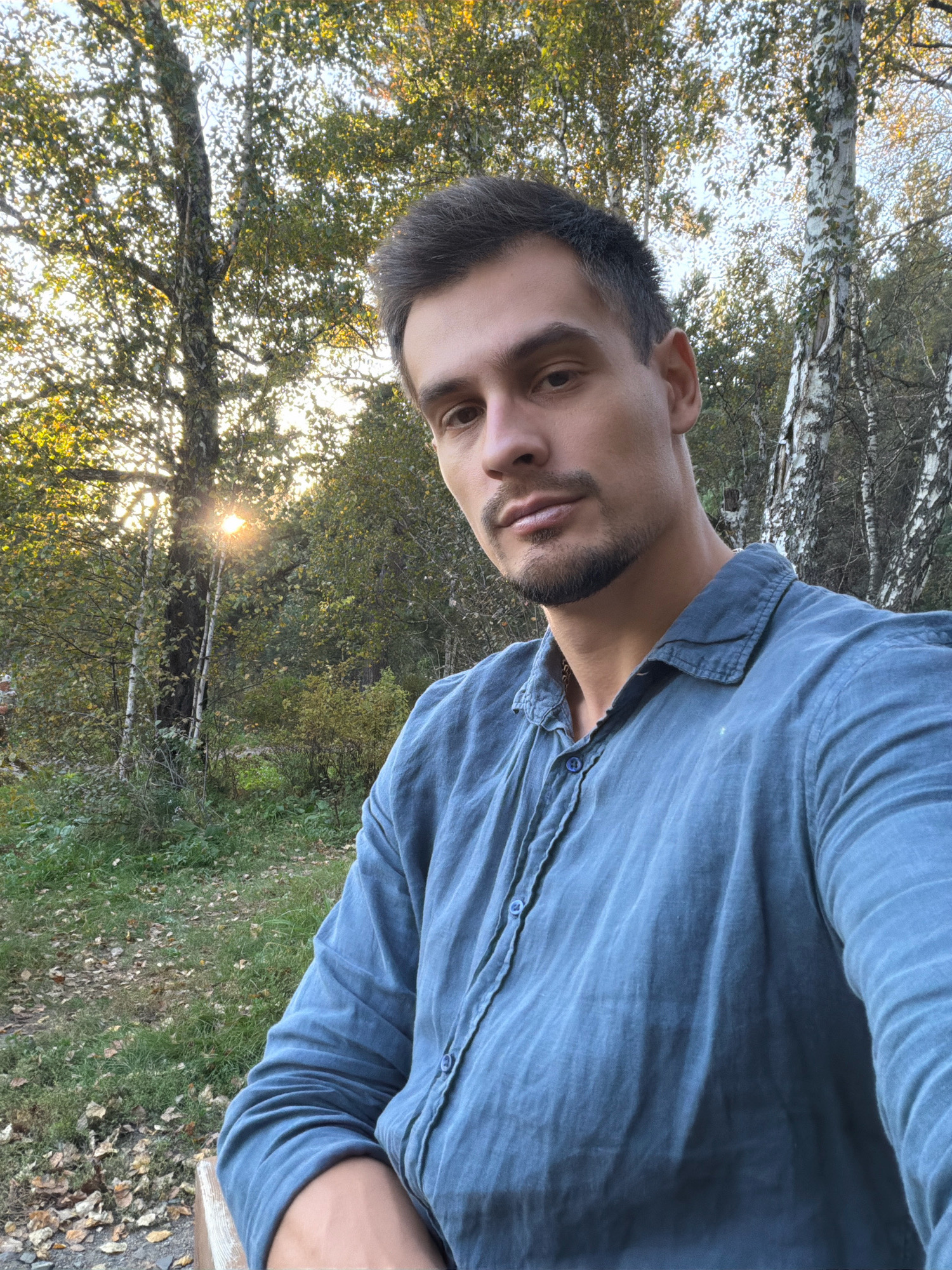 The image shows a man taking a selfie outdoors in a park-like setting, likely in Krasnoyarsk, Russia, given the presence of birch trees which are common in the region. The man is positioned in the foreground, facing the camera with a slight smile. He is wearing a blue linen shirt. The background features lush green foliage, tall trees with dappled sunlight filtering through their leaves, and a clear sky. The overall scene suggests a pleasant, natural environment, possibly during a warm season or early autumn.
