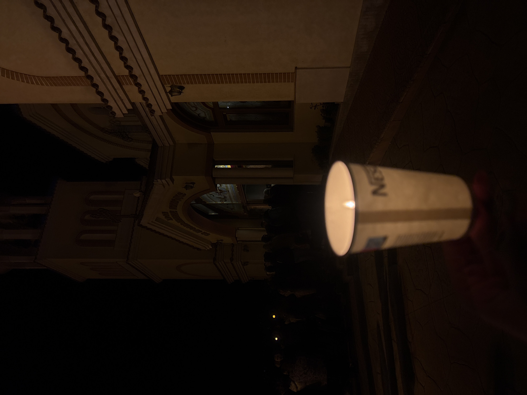 A nighttime photograph captures the facade of the Roman Catholic Cathedral of St. Louis in Plovdiv, Bulgaria. In the foreground, a hand holds a white disposable cup, possibly containing a candle, casting a glow. The building displays ornate architectural details, including arched windows and decorative trim. Below, people can be seen with candles. The light from the candles and the cup illuminate the area, while the rest of the scene fades into darkness.