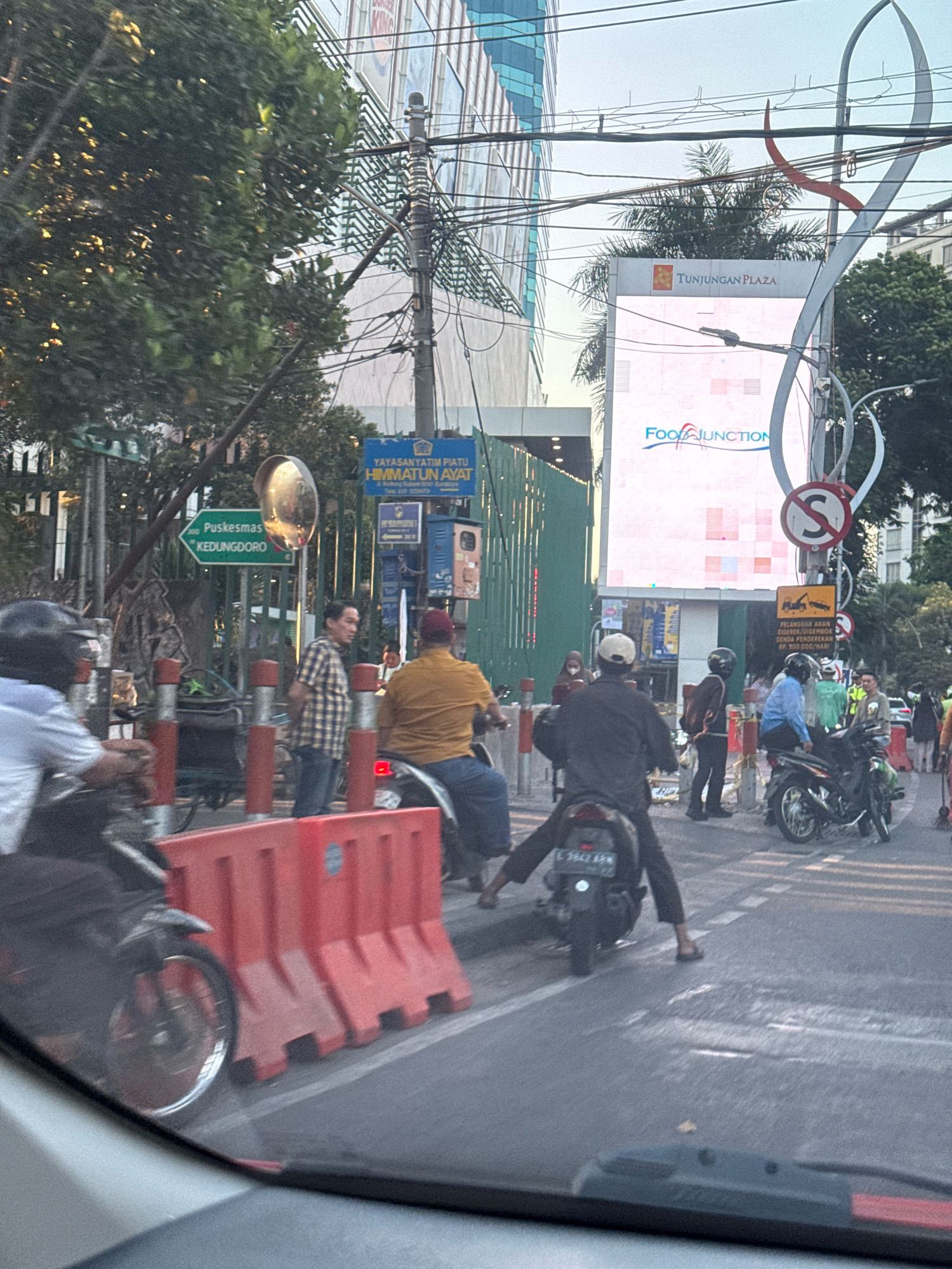 The image depicts a street scene in Surabaya, Indonesia, likely during the daytime given the lighting. In the foreground, a section of a vehicle's interior, possibly a car, is visible at the bottom. The street is lined with red plastic barriers, and several motorcycles are present. One motorcycle in the mid-ground has its license plate visible, reading "L 3847 ARM".
Several individuals are on or near motorcycles. One person in the mid-ground, wearing a black jacket and white cap, is riding a motorcycle. Another person in a yellow shirt and dark pants is also on a motorcycle, with their legs extended. Further back, a person in a plaid shirt and jeans stands near a motorcycle.
Various signs are visible in the background. One sign, predominantly blue, reads "YAYASAN YATIM PIATU HIMMATUN AYAT". Another green sign indicates "Puskesmas KEDUNG DORO". A large digital billboard in the background displays "TUNJUNGAN PLAZA" and "FOOD JUNCTION". Overhead, a complex network of electrical wires crisscrosses the scene, typical of an urban environment. Tall buildings, including what appears to be a modern skyscraper with a glass facade, dominate the skyline. The overall impression is of a busy urban intersection with ongoing traffic and infrastructure.