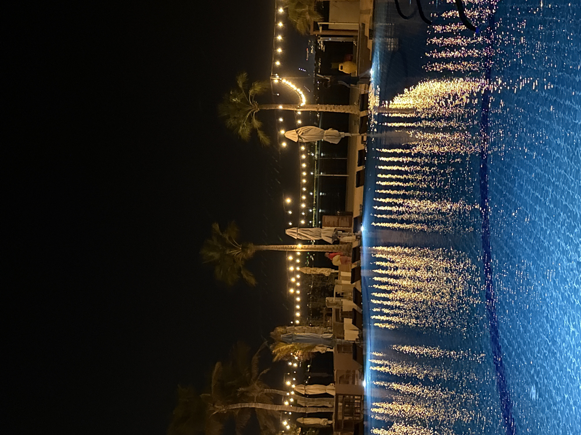 The image shows an outdoor swimming pool at night. The pool water reflects the lights from strings of bulbs hanging above, creating a shimmering effect. Palm trees and beach umbrellas are visible along the poolside, with lounge chairs arranged nearby. In the background, a cityscape can be faintly seen. One person is visible near a yellow trash can, appearing to be working. The location is likely a resort or public area in Dubai, United Arab Emirates, given the presence of palm trees and the general ambiance.