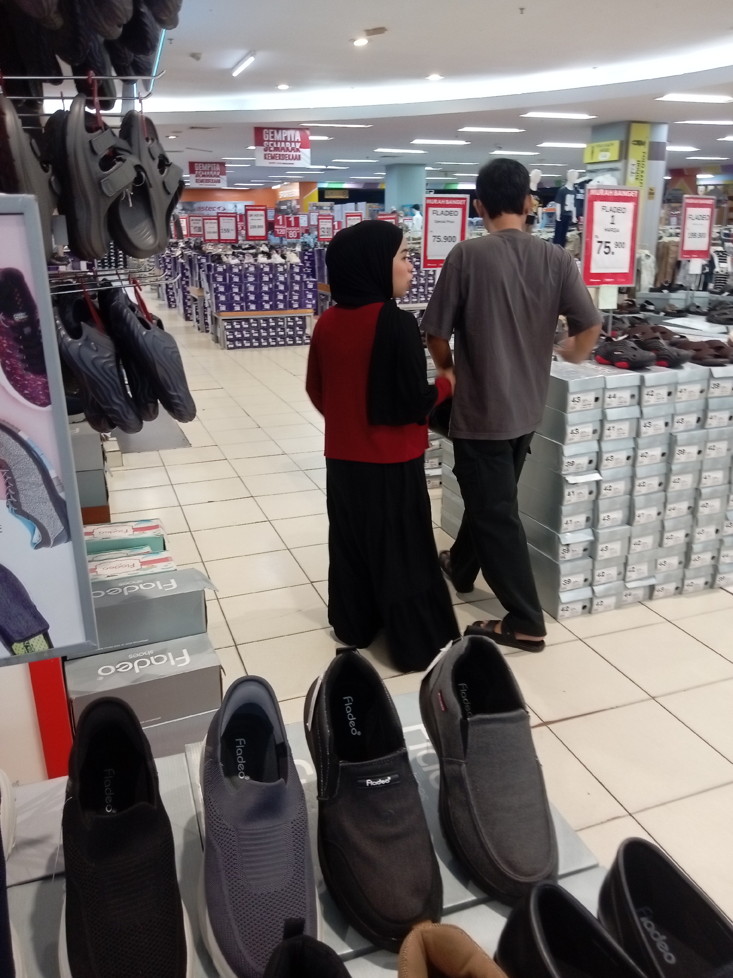 The image shows a couple browsing footwear in a retail store, likely a department store, in Batam, Indonesia. The store appears to be well-lit with fluorescent lighting and features a tiled floor. Various footwear, including sandals and shoes, are displayed on shelves, racks, and stacked in boxes. Prominent signage, some in Indonesian, indicates sales and prices, with one sign reading "MURAH BANGET" (very cheap) and another displaying "75.900." The male is wearing a grey t-shirt and dark pants, while the female is wearing a red top with a black headscarf and black pants. They are standing near a display of shoe boxes. The overall atmosphere suggests a busy shopping environment.