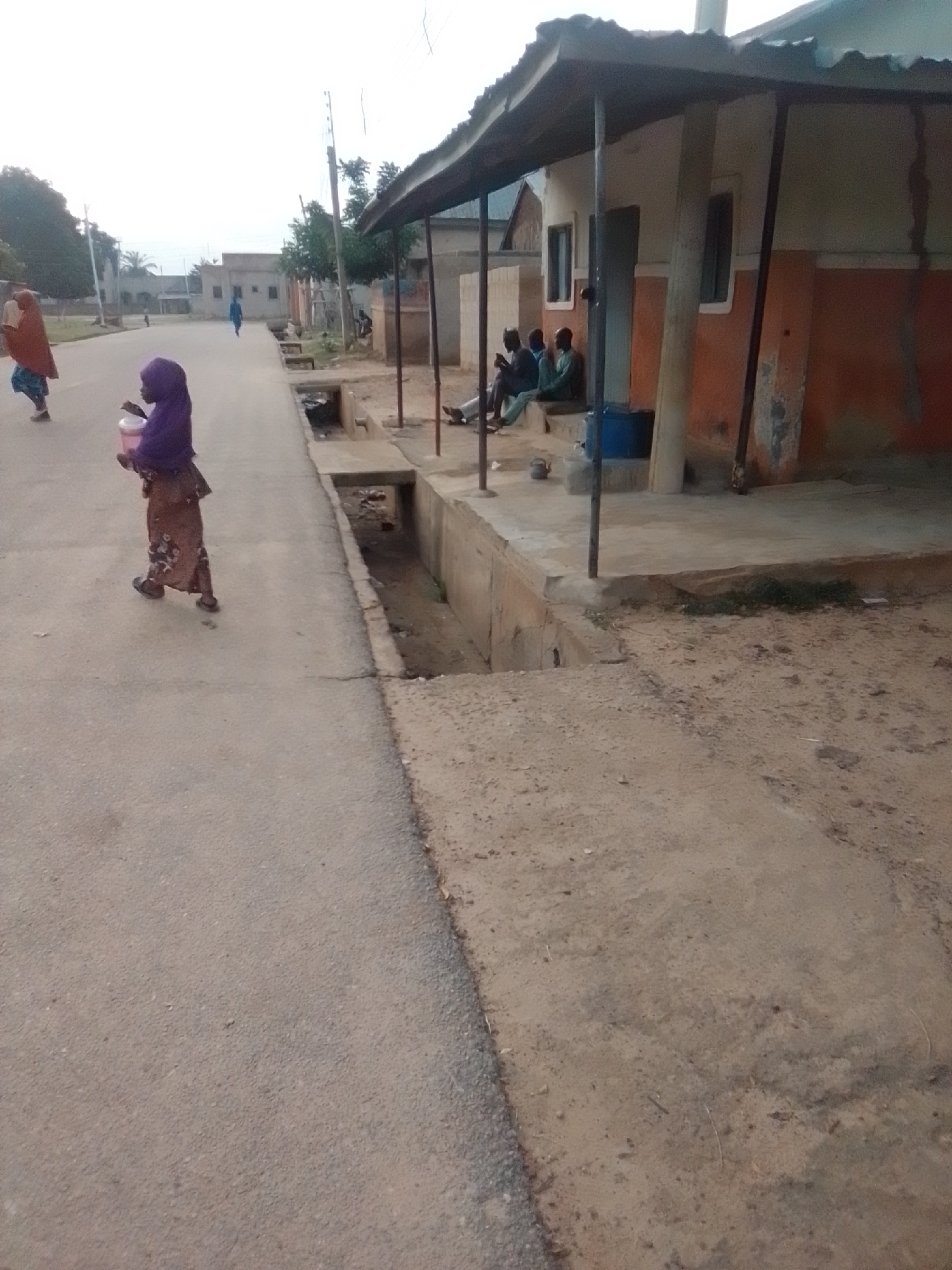 The image depicts an outdoor scene on a street in Liji Hill, Nigeria. In the foreground, a young girl wearing a purple hijab and a patterned brown dress is walking on the paved road, carrying a pink container in her right hand. To the left, another person in an orange head covering and blue patterned skirt is walking away from the camera. In the middle ground, a drainage ditch runs alongside the road, with some debris present. Adjacent to the ditch, a building with an orange and white facade is visible, featuring a covered porch area where three individuals are seated. A blue plastic barrel is also present near the porch. The background shows more buildings and vegetation under an overcast sky.