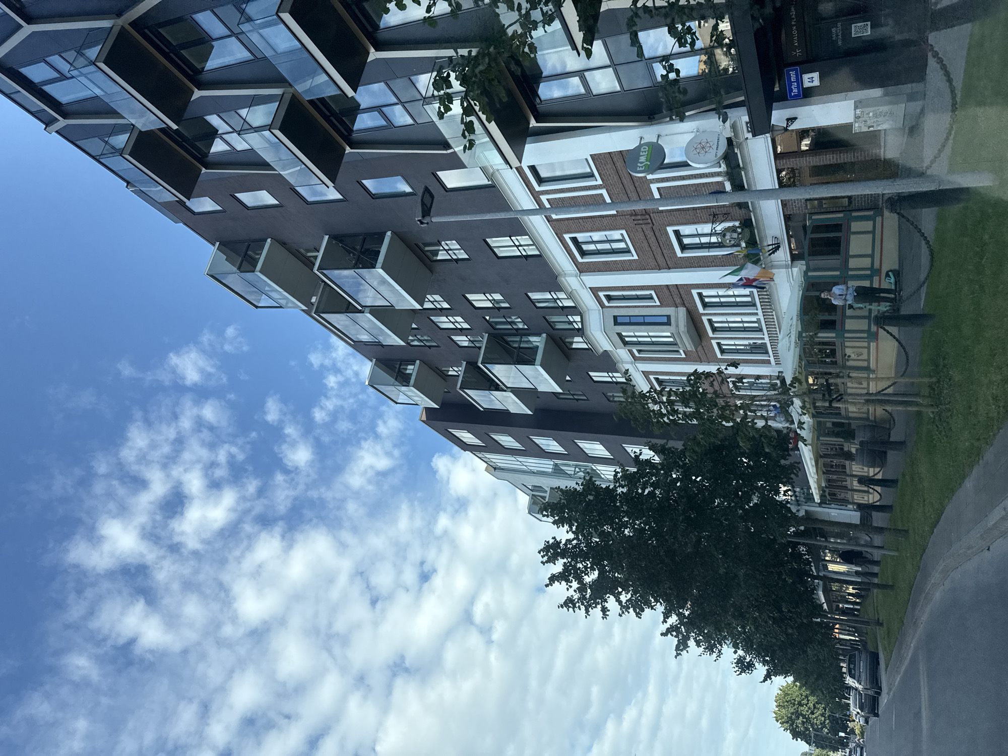 The image captures a street view in Tallinn, Estonia, specifically along Tartu mnt. A modern, dark-colored apartment building with prominent glass balconies dominates the right side of the frame. The building features a lower section with red brickwork and white trim, housing commercial spaces. A sign with "ESMED" and a circular logo is visible on this lower section. To the left of the building, a row of trees lines a sidewalk and a street with parked cars. The sky is blue with scattered white clouds. A person in a light blue shirt and dark pants stands on the sidewalk near an entrance to the building. In the background, other vehicles are visible on the street. The scene is set on a clear day.