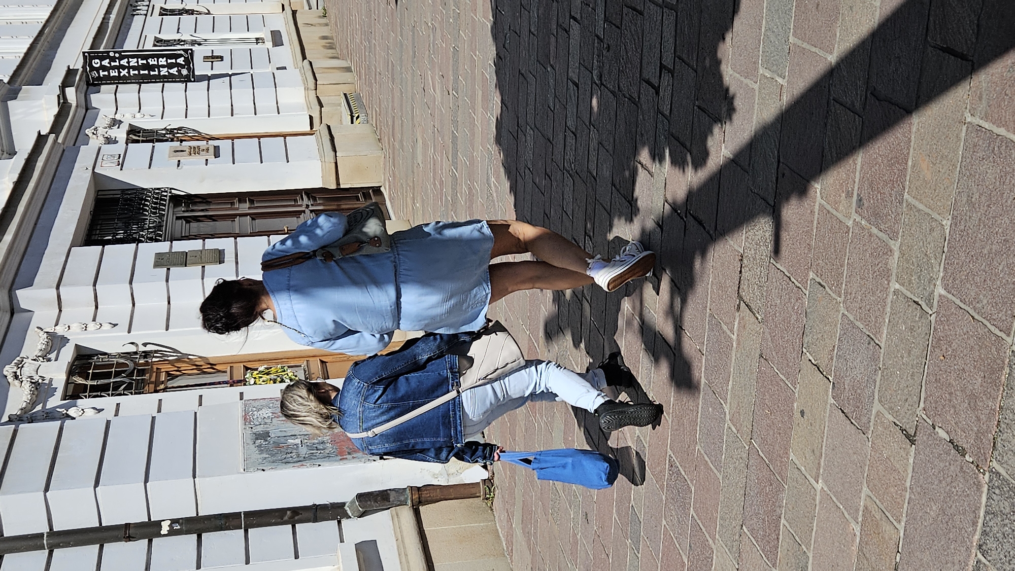 Two individuals are walking away from the camera on a cobblestone sidewalk in front of a light-colored building. The building has decorative architectural elements, windows with bars, and signage. The individual on the left is wearing a denim jacket, light-colored pants, and sneakers, and is carrying a blue tote bag. The individual on the right is wearing a light blue dress and white sneakers, and is carrying a brown bag over their shoulder. The scene is outdoors with strong sunlight casting shadows on the ground. The signage on the building, "GALANTÉRIA TEXTIL," identifies the location as Galantéria Textil in Prešov, Slovakia.
