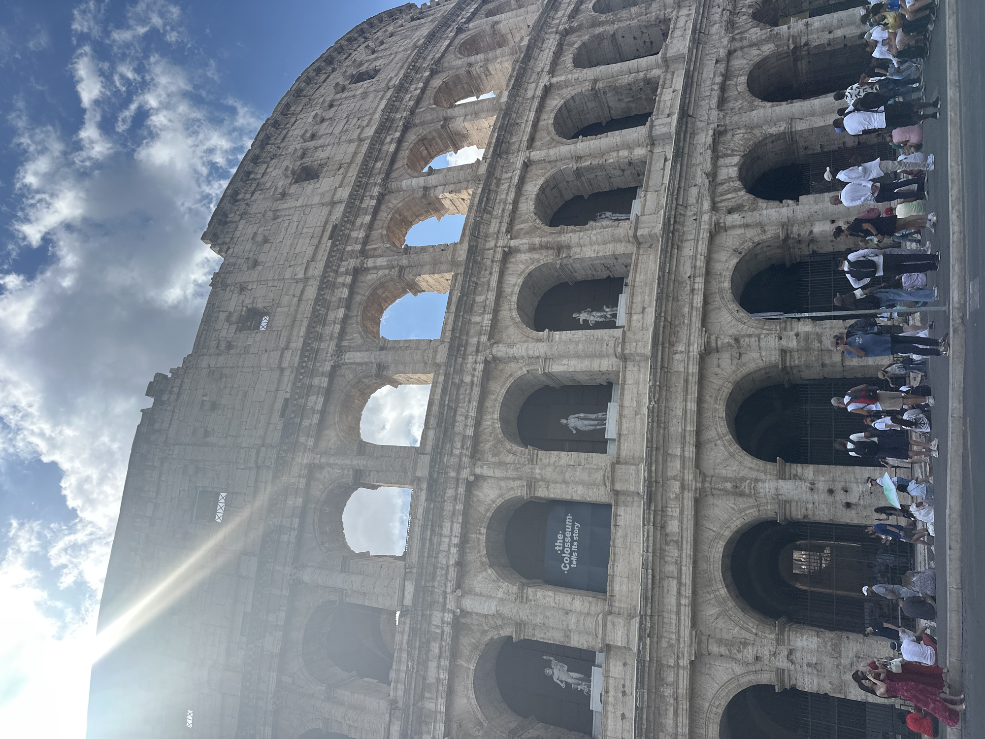 The image shows a low-angle, wide shot of the Colosseum in Rome, Italy. The sun is shining brightly from the upper left, casting rays across the ancient stone structure. The Colosseum is a massive oval amphitheater with multiple tiers of arches. In the foreground, a crowd of tourists is gathered, with some sitting and others standing. They are diverse in age and attire, and many are looking at the Colosseum or taking pictures. A sign within one of the arches reads, ".the. -Colosseum- tells its story". The sky above is a mix of blue with scattered white clouds.