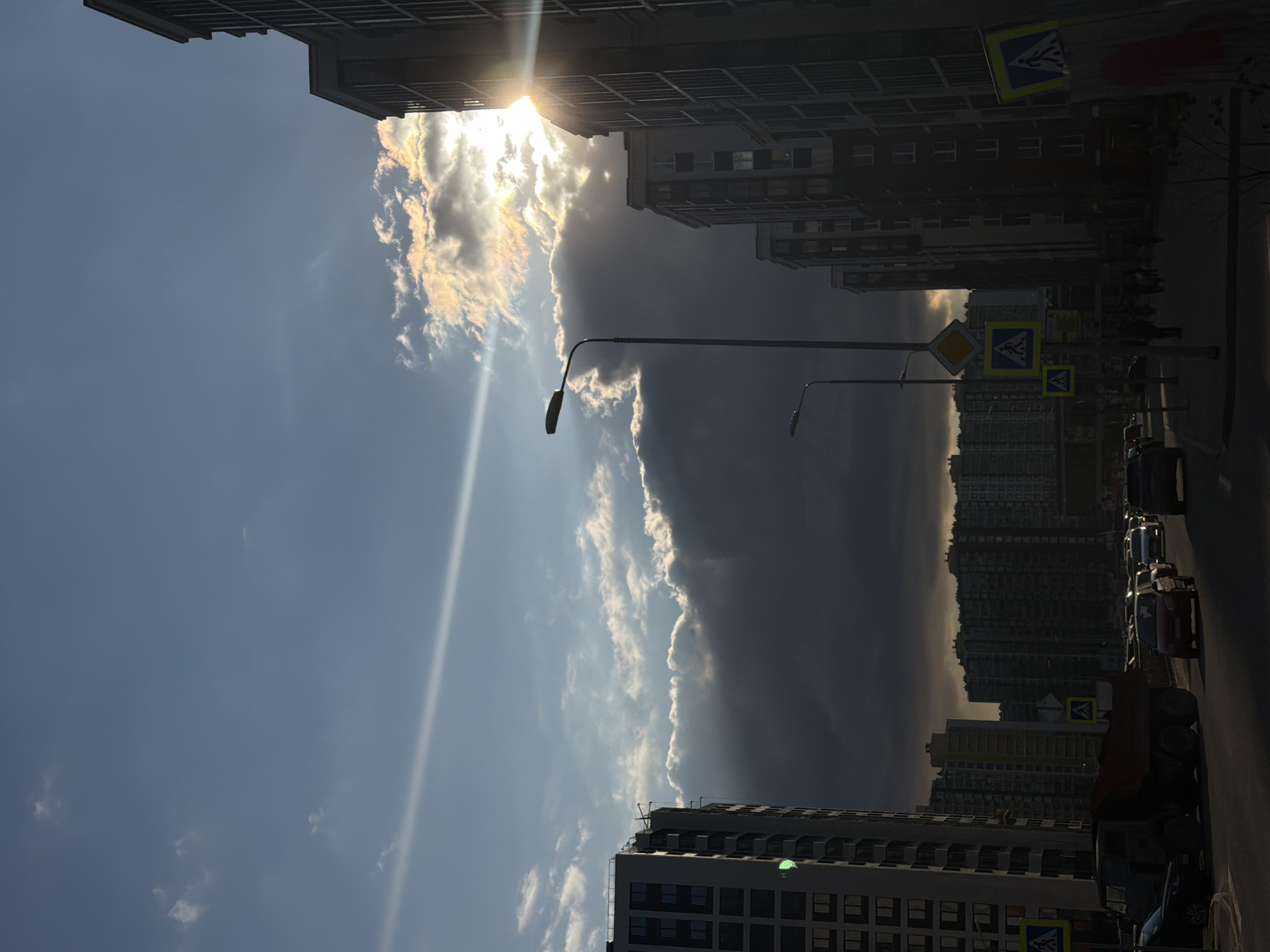 The image captures a street in Murino, Russia, lined with multi-story residential buildings. The scene is dominated by the sky, where sunlight breaks through a dramatic cloud formation, creating a stark contrast between light and shadow. Streetlights are visible along the road, and pedestrian crossing signs are present. Several cars and what appears to be construction vehicles are parked along the street. Individuals appear as silhouettes along the street. The image is taken from a low angle, emphasizing the height of the buildings and the sky.