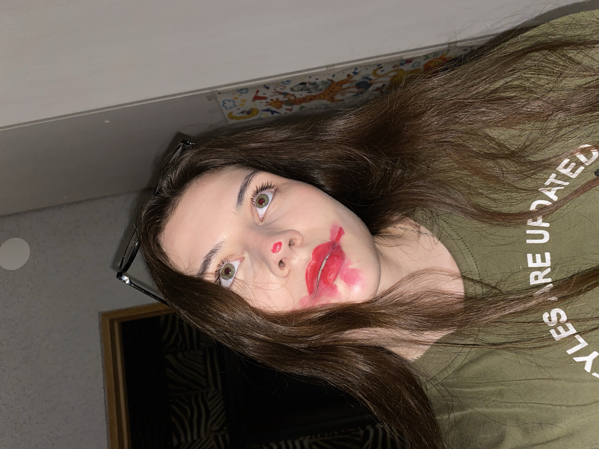 A close-up, rotated portrait shows a young person with long, wavy dark brown hair sprawled out, looking upwards with wide, light green eyes. The person's face is stained with bright red lipstick, smudged around the mouth, chin, and with two distinct red dots on the nose, giving a playful or mischievous appearance. Their eyebrows are dark and well-defined.
They are wearing an olive green t-shirt, on which white text is partially visible, reading "YLES ARE UPDATED". A thin silver necklace chain is visible around their neck. Black-framed glasses are tucked into their hair near the left ear.
The background features various elements suggesting an indoor setting. Behind the person's head, a light grey textured wall with a circular white light fixture or decor is visible. Further back and above, a lighter wall or ceiling transitions into a colorful patterned border adorned with cartoonish motifs including a giraffe, clouds, and stars. In the lower left corner of the frame, a wooden picture frame with a black and white striped or patterned interior is visible. The lighting is bright and even, indicating an indoor environment, possibly with artificial light.