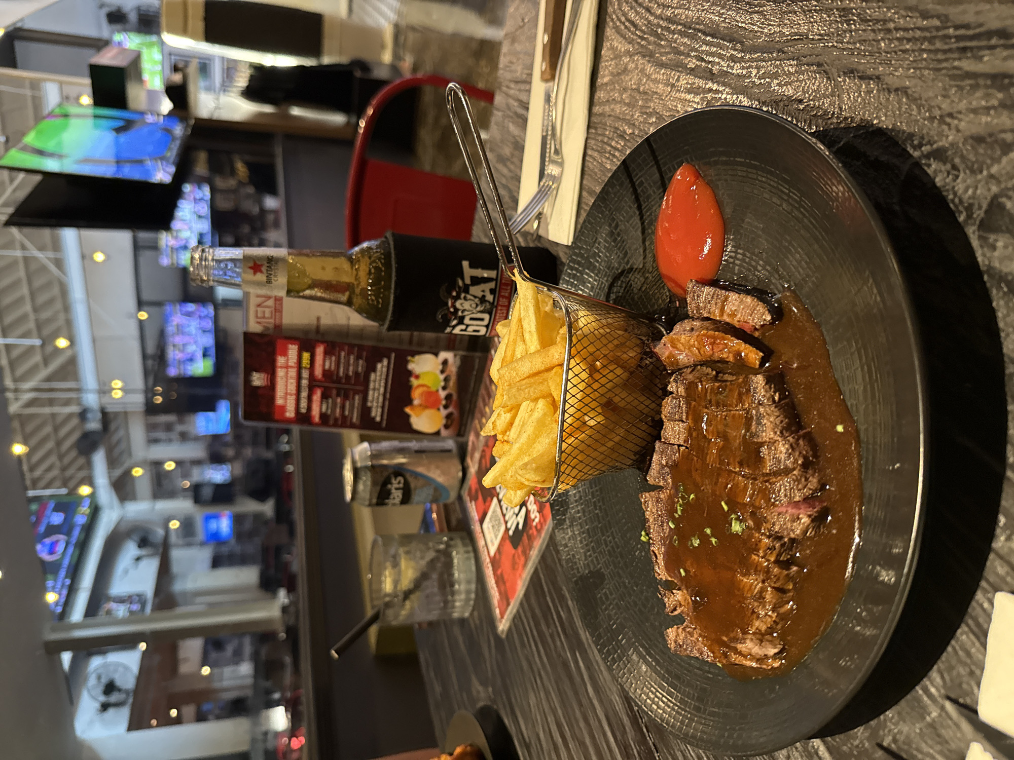 This indoor image captures a casual dining experience, likely during the evening or night, in a restaurant or bar in Kuta, Indonesia. The primary focus is on a meal served on a dark, textured tabletop.
On a black, ridged plate, sliced steak, generously coated in a dark brown sauce and garnished with chives, is presented next to a small metal basket filled with golden french fries. A prominent dollop of bright red sauce, likely ketchup, is also on the plate. Accompanying the food are several beverages: a "Bintang Pilsener" beer bottle, partially labeled "BINTANG CRISTAL", nestled in a black koozie emblazoned with a goat logo and the text "GOAT BEST OF ALL TIME". Also on the table are a clear glass with ice and a dark straw, and a partially visible "Polaris" branded can. A menu or promotional leaflet, displaying text such as "MEN", "SHOT", "PROMOTION", "CLASSIC COCKTAIL", and "BUY 1 GET 1", lies beside the drinks. A fork and knife, wrapped in a white napkin, are partially visible to the right of the plate.
The blurred background reveals the interior of the establishment, characterized by multiple television screens displaying indistinct images, likely sports or entertainment. Overhead lights create a warm ambiance, and reflections are visible on shiny surfaces. A red chair and various architectural elements are discernible, suggesting a spacious venue. While no specific individuals are clearly identifiable, the setting indicates a lively, relaxed atmosphere where patrons are enjoying food and drinks, consistent with a pub or casual restaurant. The presence of Bintang beer is a strong location cue for Indonesia.