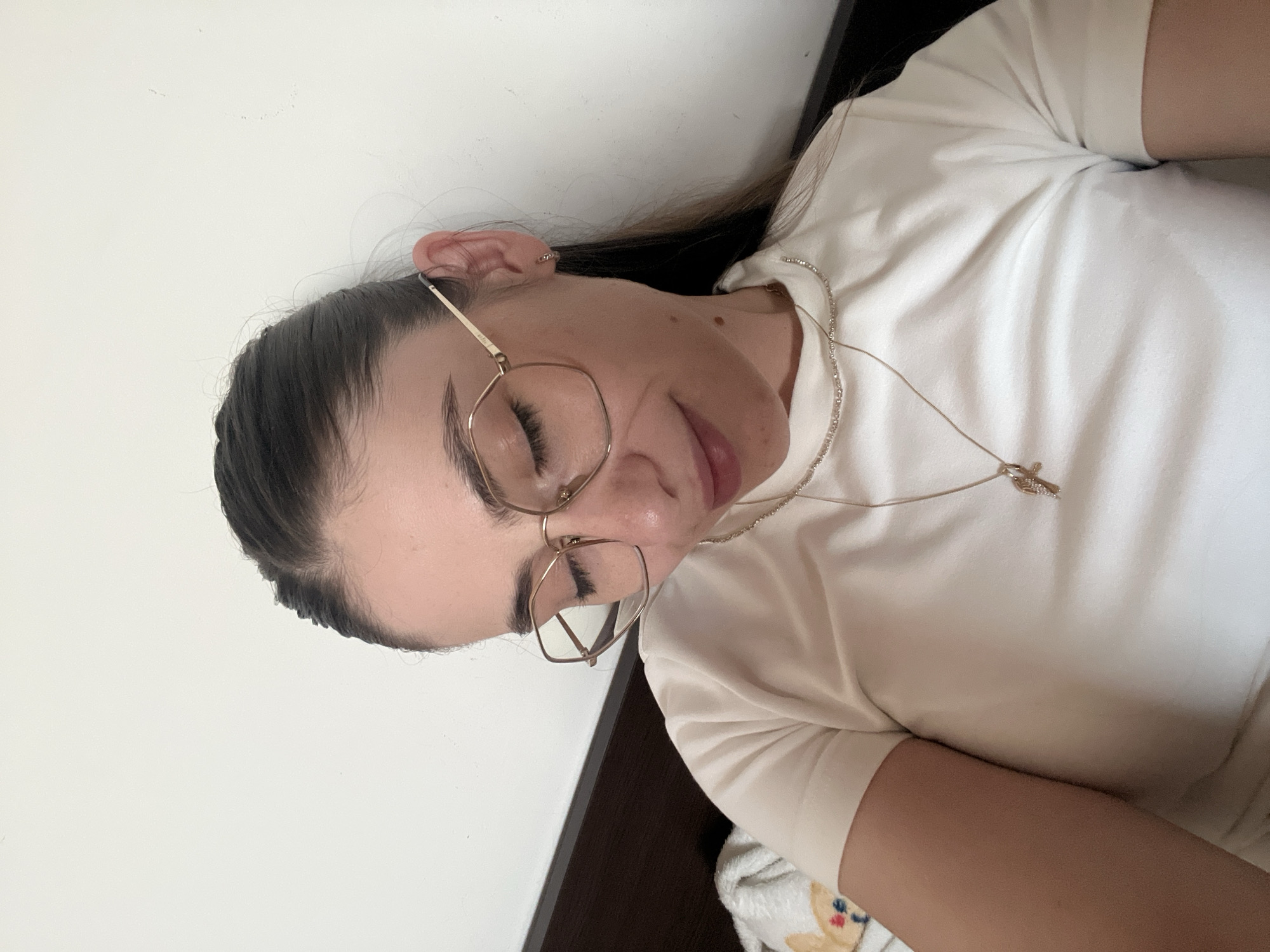 A young woman with her eyes closed is pictured from the chest up, leaning back against a white wall. She is wearing glasses with gold frames and a white t-shirt. A thin gold necklace with a cross pendant rests on her shirt. Her dark hair is pulled back in a braid. The woman is wearing small gold hoop earrings. The scene appears to be indoors, possibly in a bedroom, with a dark wooden headboard visible to the left of the woman.