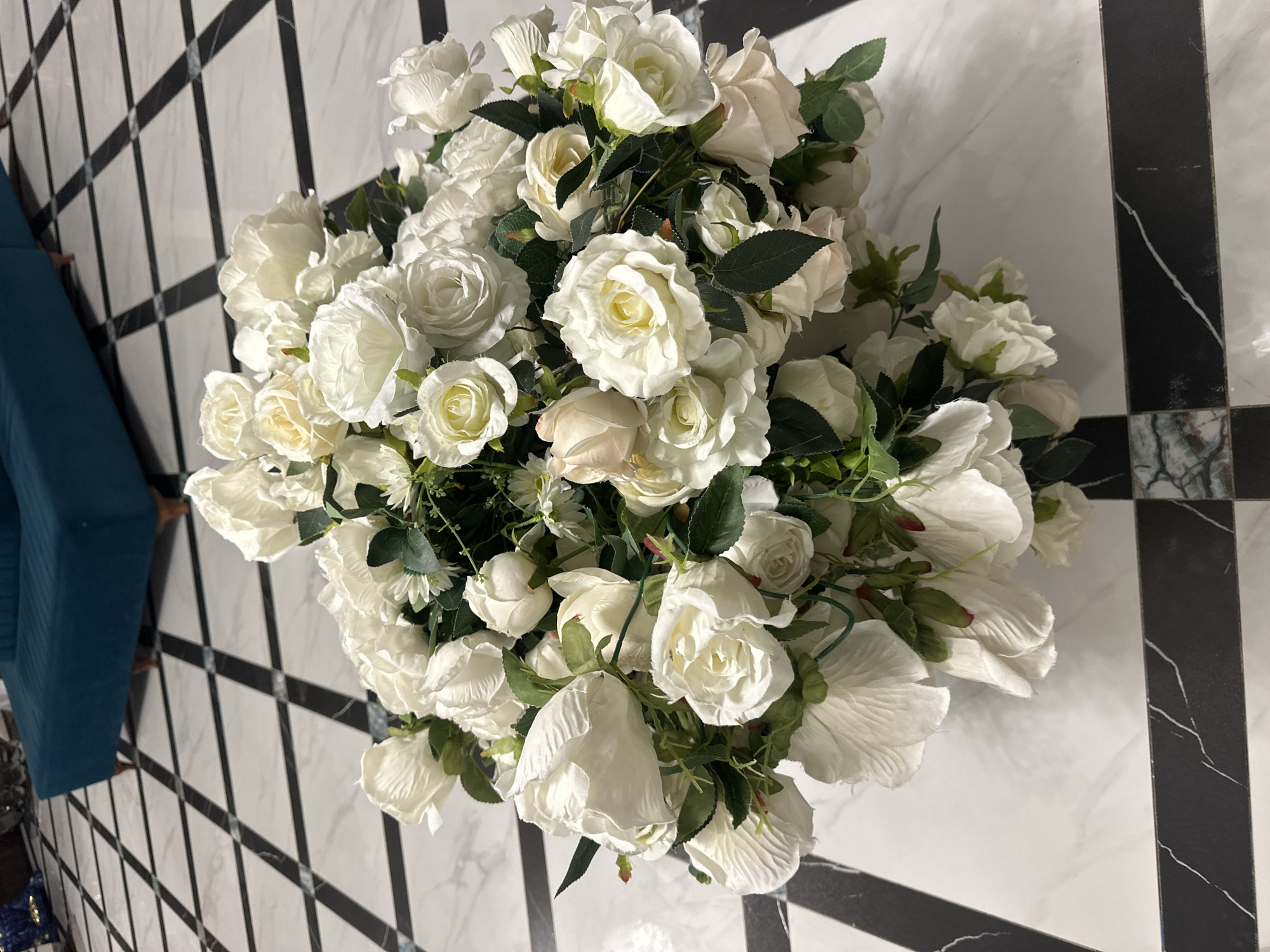 An overhead, medium shot captures a lush bouquet of artificial white and cream roses and greenery. The flowers are densely packed, with some buds and fully bloomed roses showing. The bouquet rests on a white marble floor with a striking black and white checkered pattern. In the background, a portion of a teal upholstered sofa is visible. The lighting is bright, casting soft shadows from the bouquet onto the floor. This scene appears to be an indoor setting, likely a residential or commercial space, in Bishkek City, Kyrgyzstan.