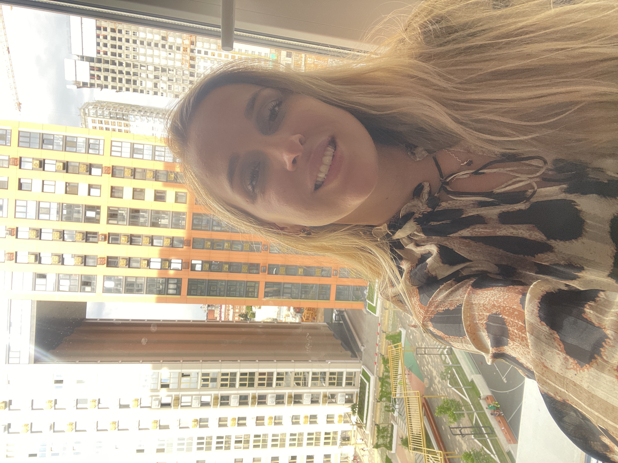 A selfie-style photograph depicts a blonde woman smiling at the camera. She is wearing a brown and black patterned top. The background shows a cityscape, likely taken from a high-rise building. The buildings in the background are multi-story, with various colors (orange, white) and architectural styles. There is construction visible in the background. The image is rotated 90 degrees counter-clockwise. There is no information present in the image to confirm the location of Perm, Russia.