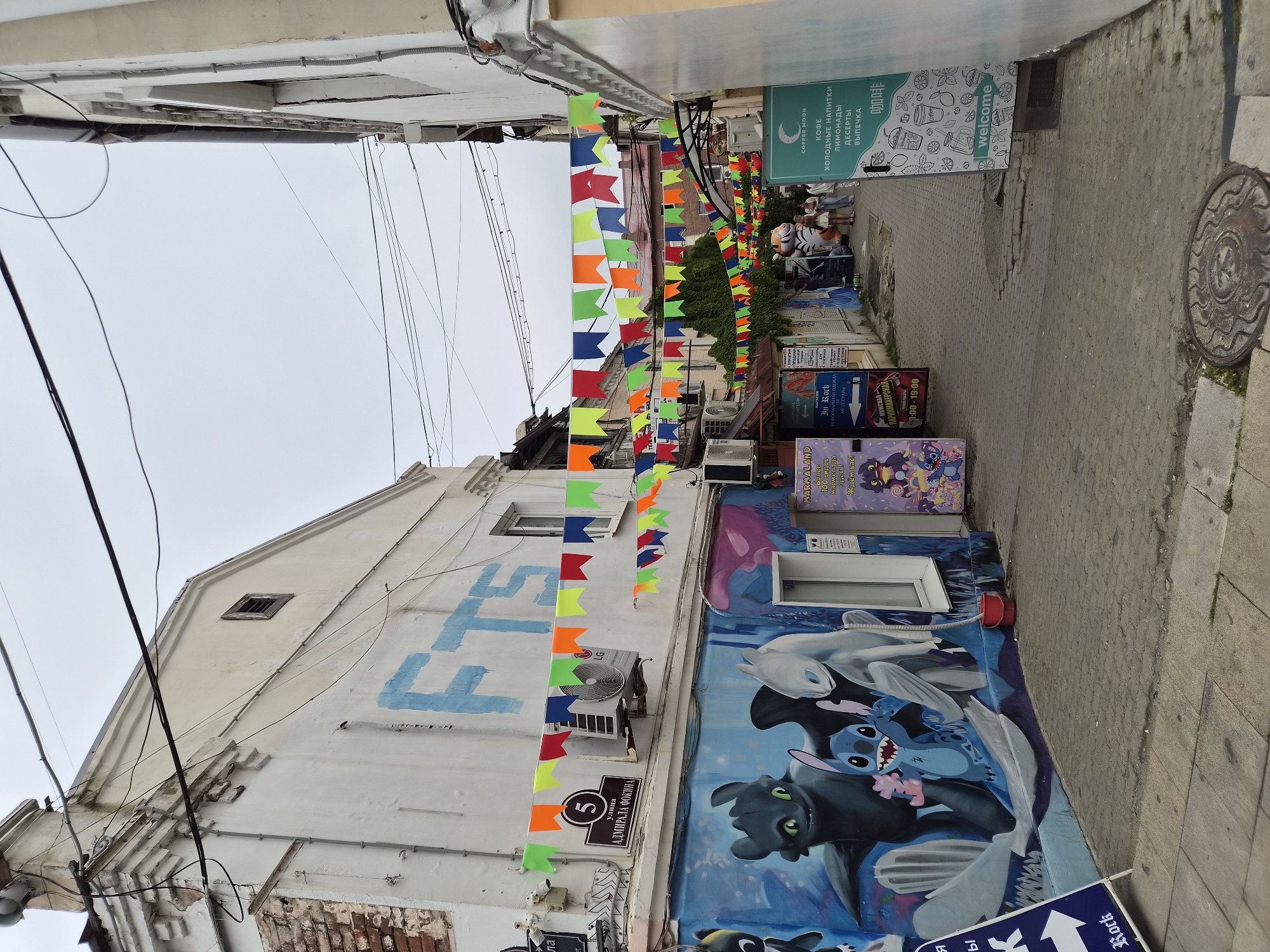 This image captures a narrow pedestrian alleyway in Vladivostok, Russia, adorned with vibrant, multi-colored pennant banners strung overhead. The buildings lining the alley are old and show signs of wear, with peeling paint and exposed brickwork on the left. The walls are decorated with large, colorful murals, including a depiction of characters from "How to Train Your Dragon" and Stitch from "Lilo & Stitch" on the left.
Several businesses are visible along the alley. On the left, a sign indicates "In Rock," a shop selling musical instruments, and further down, a "COFFEE MOON" sign advertises coffee, lemonades, desserts, and pastries. Small shops and kiosks are nestled between the buildings, and some have their storefronts open, revealing goods and displays within.
An air conditioning unit is mounted on the wall of the building on the left, beneath a street sign that reads "ул. Адмирала Фокина" (Admiral Fokin Street). The ground is paved with cobblestones, and a manhole cover is visible in the foreground. A few people are walking through the alley, appearing to be tourists or shoppers. The overall atmosphere is lively and colorful, despite the aged architecture. The sky is overcast.