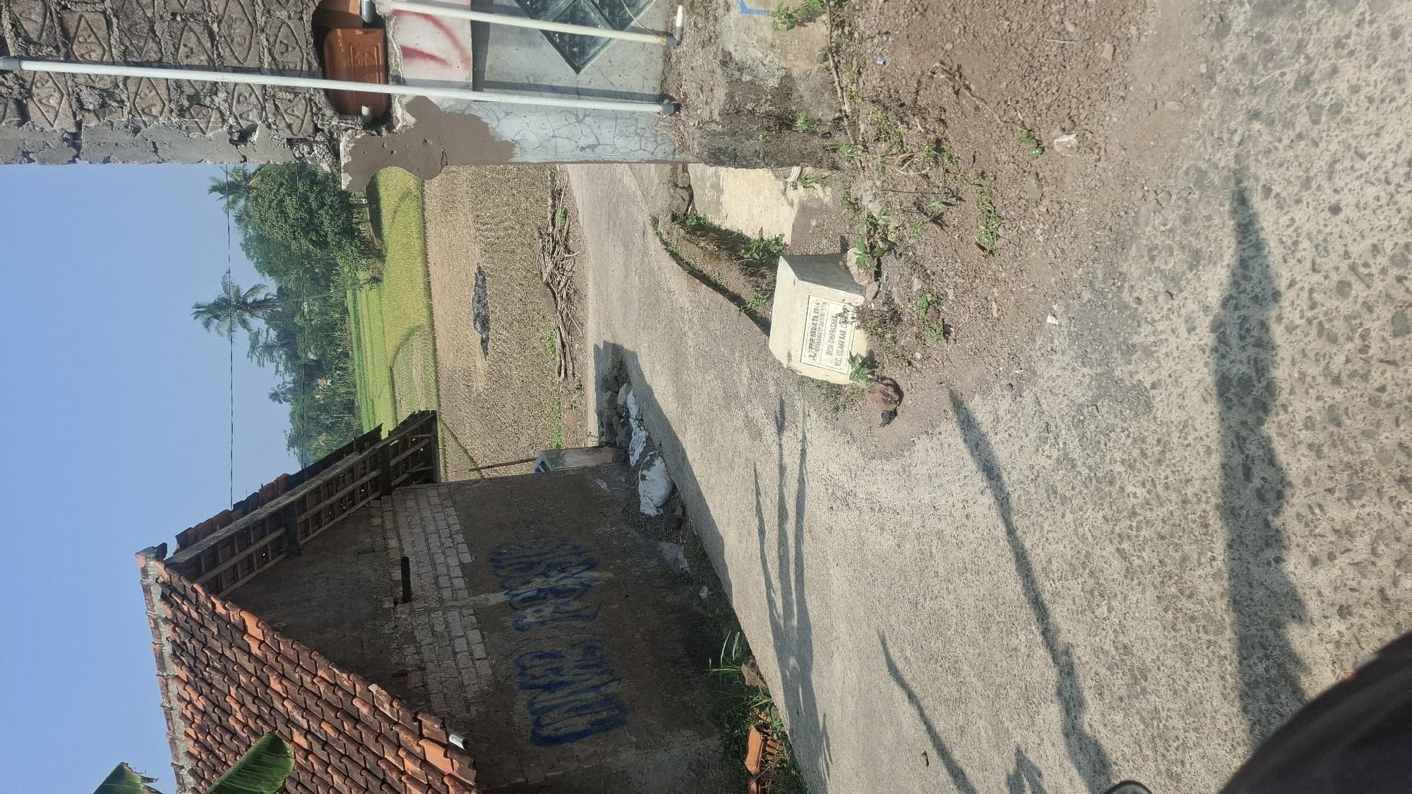 The image depicts a rural scene in the city of Cianjur, Indonesia, under a clear blue sky. In the foreground, a paved road curves through the scene, casting long shadows from an unseen object to the left. To the left of the road, a dilapidated brick building with a tiled roof stands partially in shadow. Blue graffiti adorns the side of this building. Further down the road, a small concrete marker with inscriptions is visible. To the right of the road, a rough concrete wall with visible rebar and pipes rises up. Beyond these structures, a lush green rice paddy stretches out, dotted with palm trees and other vegetation. The overall environment suggests a developing or rural area within Cianjur. There are no people visible in the image.