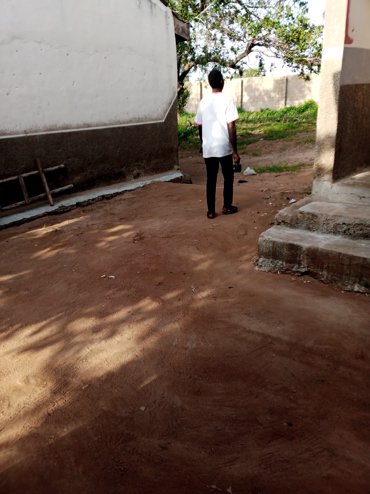 A person, seen from behind, stands in an outdoor area with reddish-brown dirt ground. They are wearing a white t-shirt with red trim on the collar and sleeves, and black pants. The person is holding a dark object, possibly a camera or phone, in their right hand. To the left, a section of a building with a textured, dark brown base and a lighter, rough-textured upper wall is visible. A wooden ladder-like structure is propped against the base of this building. To the right, there are concrete steps leading up to another building. In the background, a light-colored brick wall borders an area with green grass and trees. The scene appears to be an outdoor courtyard or alleyway.