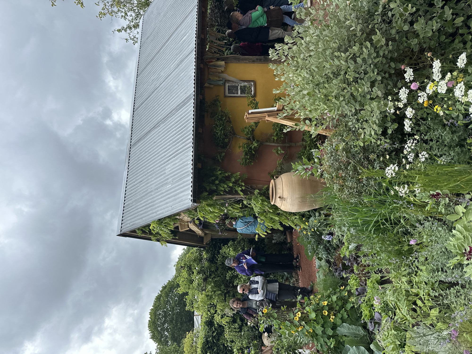 The image depicts a garden scene in London, United Kingdom. A group of people stand in a lush garden adjacent to a building with a corrugated metal roof. The building has a yellow facade with an orange base, a small window, and is decorated with climbing plants. An easel stands near the building, suggesting artistic activity. In the garden, there is a large ceramic pot and a diverse range of plants, including flowering varieties. The sky is overcast, with cloudy weather conditions. People are standing on a path in the garden, some interacting with each other.