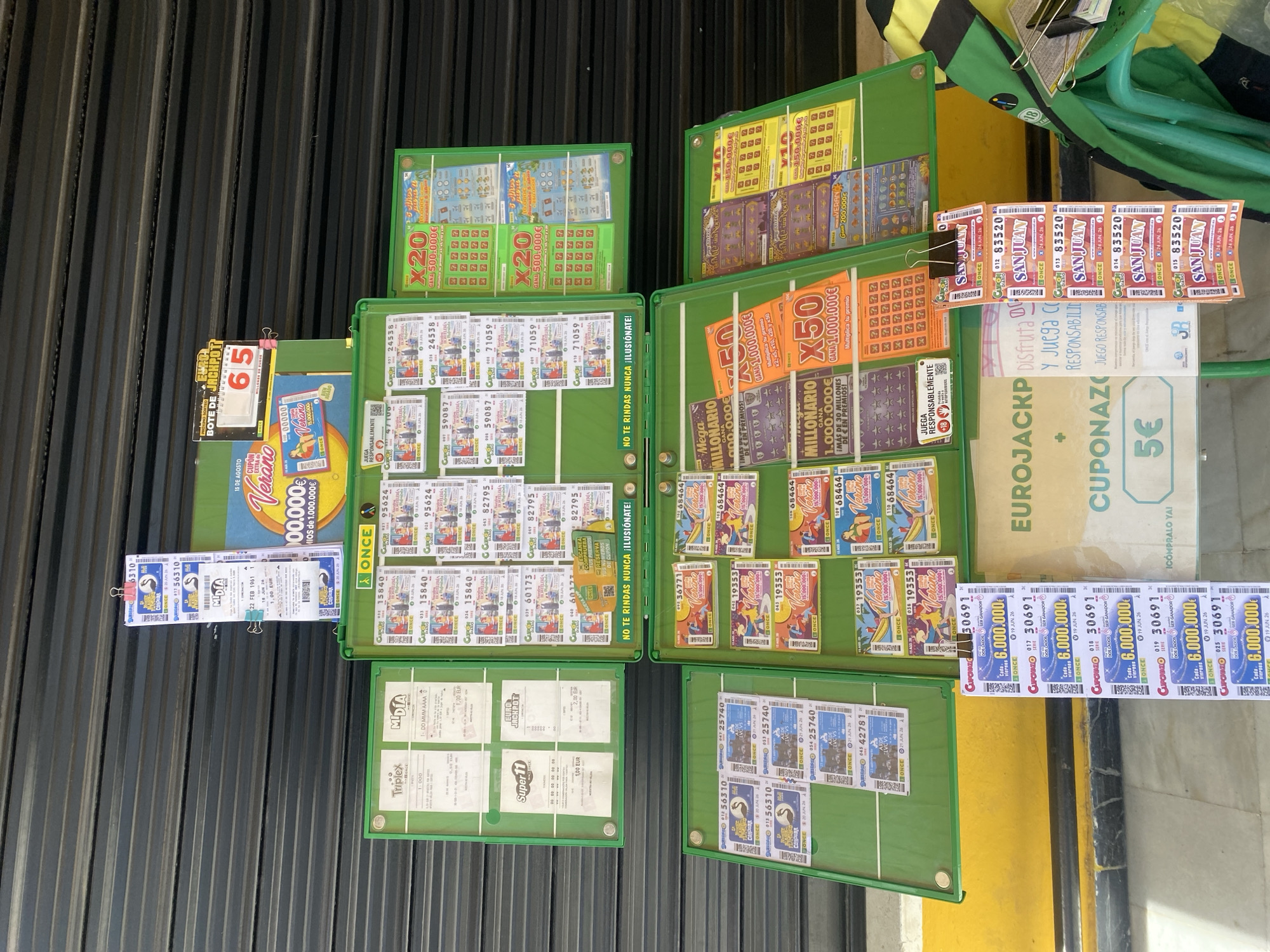 The image shows a close-up, top-down view of a lottery stand in Bel Air, Spain. The stand is filled with various lottery tickets, primarily from "ONCE" (Organización Nacional de Ciegos Españoles), a Spanish lottery and social enterprise. The tickets are displayed in green plastic trays and some are clipped together.
The subjects are primarily the lottery tickets themselves, featuring vibrant colors and different prize amounts like "GANA 500.000€," "GANA 1.000.000€," and "6.000.000€." There are also promotional banners and signs. One prominent sign reads "BOTE DE JACKPOT 100.000€ Verano," indicating a jackpot prize for a summer lottery. Another banner advertises "EURO JACKPOT" with a prize of "5€." Text on the tickets includes dates like "20 JUN 26" and "24 JUN 26," suggesting these are for upcoming draws. Some tickets are labeled "MILLONARIO," indicating a millionaire prize.
The activity taking place is the sale and display of lottery tickets, likely at an outdoor stall given the presence of a dark, corrugated metal surface in the background, possibly a storefront or stall facade. The lighting suggests it could be daytime, with natural light illuminating the brightly colored tickets. The overall impression is one of a busy point of sale for chance-based games. The text "NO TE RINDAS NUNCA ¡ILUSIÓNATE!" (Never give up, be hopeful!) and "JUEGA RESPONSABLEMENTE" (Play responsibly) appear on some tickets, reinforcing the theme of hope and responsible gambling.