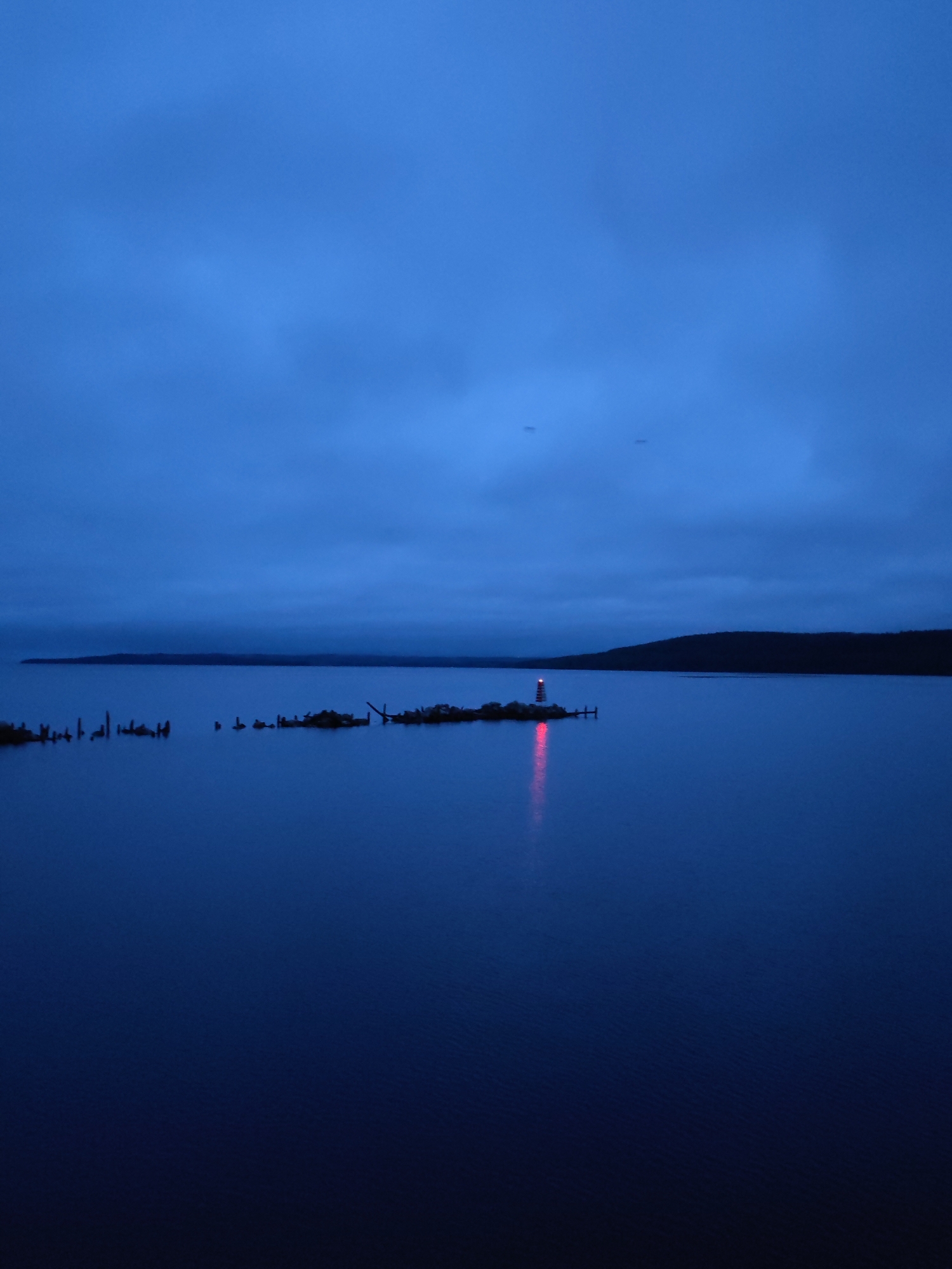 The image captures a serene, low-light waterscape, likely a lake or large bay near Medvezhyegorsk, Russia, possibly Lake Onega. The scene is set during either dusk or dawn, characterized by a dominant deep blue hue across the sky and water.
The primary subject is a small lighthouse situated on a rocky island or peninsula in the mid-ground. The lighthouse emits a vivid red light, which casts a prominent crimson reflection streak across the calm surface of the water. Surrounding the lighthouse on its small landmass are various rocks and what appear to be old, dilapidated wooden pilings or remnants of structures. To the left of the lighthouse island, more scattered wooden posts or stumps protrude from the water, suggesting submerged structures or shallow areas.
The expansive sky above is a uniform deep blue, covered with soft, indistinct clouds. Two very faint, tiny specks are barely discernible in the upper-mid section of the sky, whose nature is ambiguous (possibly distant birds or aircraft). The water itself is dark blue and largely smooth, reflecting the sky and the lighthouse's glow, indicating calm weather conditions. The distant horizon reveals a dark, undulating landmass, likely a forested shoreline or hills.
No people or direct activity are visible, lending a tranquil and somewhat isolated atmosphere to the scene. The prominent red light of the lighthouse serves as a navigational beacon in the subdued lighting. There is no visible text in the image.