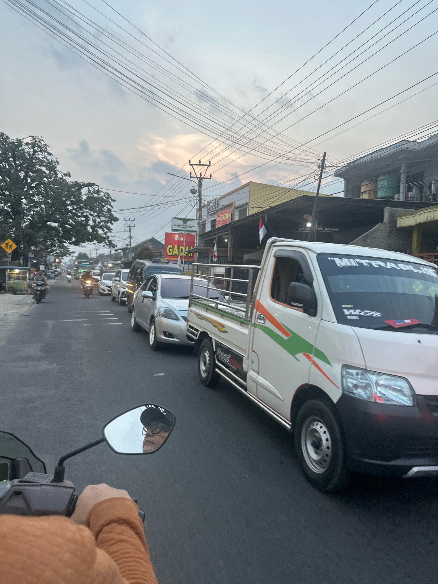 The image captures a street scene in Saruni, Indonesia, characterized by moderate traffic and overcast skies. In the foreground, a person's hand in an orange sweater is visible, holding the handlebars of a motorcycle. The motorcycle's rearview mirror reflects the rider, who is wearing glasses and a cap, along with the sky.
Dominating the right side of the frame is a white pickup truck with green and orange decals. Its side displays the text "MITRAGUIS" and "WRZS TRON". The truck is part of a line of vehicles on the road. Behind it, a silver car is clearly visible, followed by a dark-colored van and several other cars and motorcycles.
On the left side of the street, trees line the sidewalk, and several buildings with signage are present. One sign, in red and yellow, reads "GADAH". Another sign depicts two figures walking with backpacks, indicating a pedestrian crossing. Streetlights and utility poles with numerous wires crisscross the sky. The overall atmosphere suggests a typical day in a moderately busy urban environment, possibly during the late afternoon or early evening given the lighting.