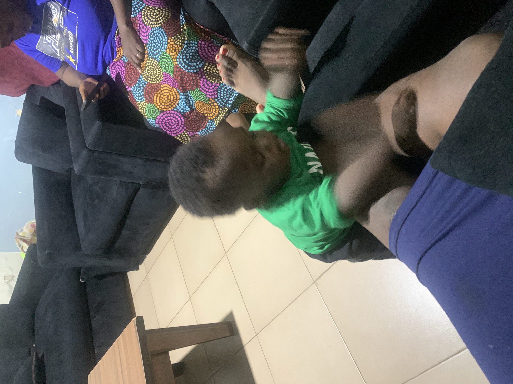 The image shows a top-down perspective of a baby lying on a tiled floor next to a black sofa and a wooden table. The baby is wearing a bright green long-sleeved shirt and appears to be reaching out. Nearby, a person's bare foot with red nail polish is visible, along with a patterned fabric that appears to be part of clothing or a blanket, featuring circles of various colors like pink, orange, blue, and yellow. Another person is partially visible in the background wearing a blue t-shirt with text on it, and holding a dark object, possibly a phone. The scene is indoors, with tiled flooring and a wall visible in the background. The lighting suggests it could be daytime or evening, and there are no obvious weather cues. The overall impression is a casual, domestic setting, possibly a living room, with a focus on the baby's playful activity. There is some text visible on the blue t-shirt in the background, but it is partially obscured and difficult to read.