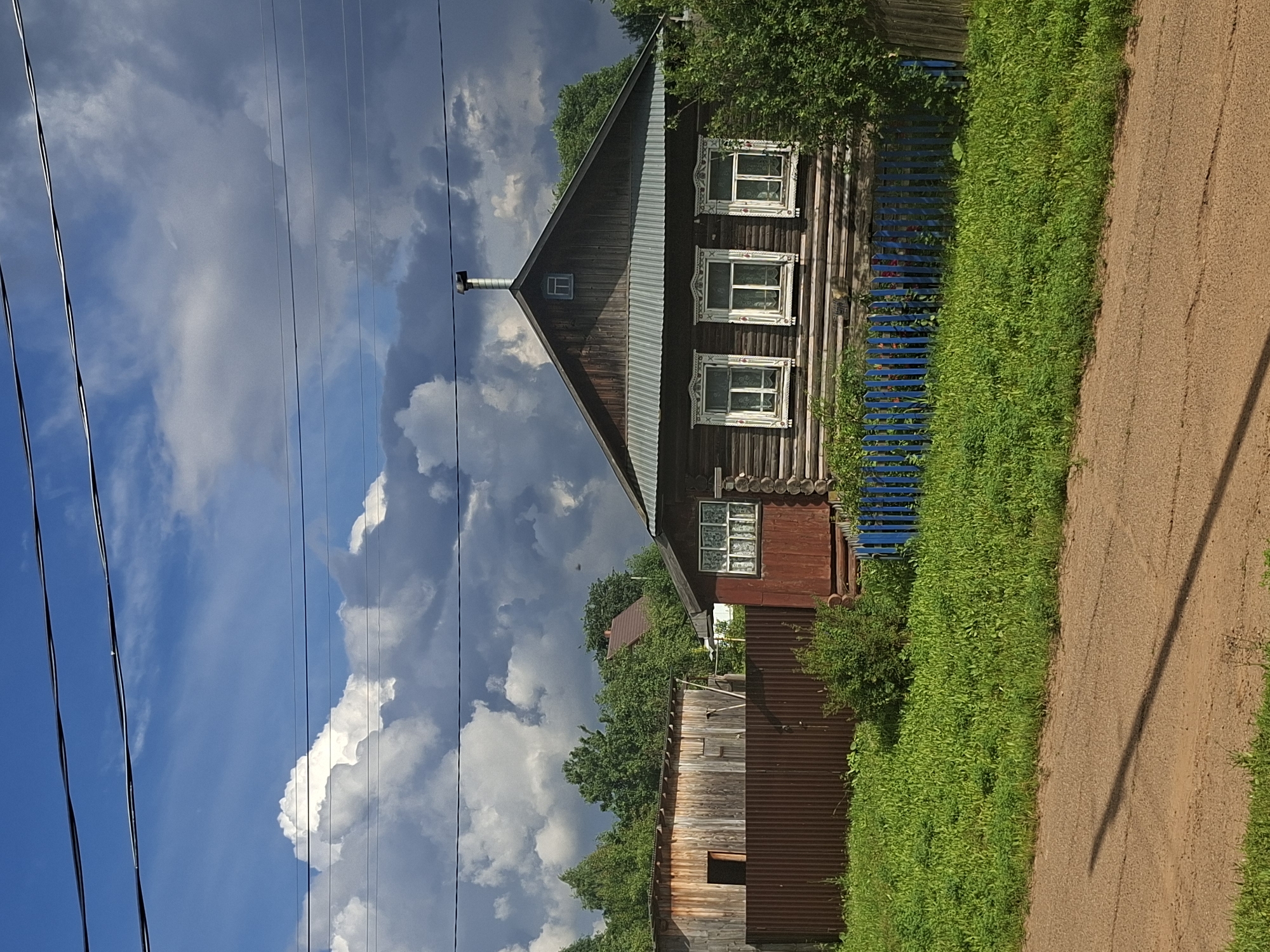 This media file depicts a rural scene in Karsha, Russia. The primary subject is a traditional wooden house with an angled roof and a prominent chimney. Three windows with decorative white trim are visible on the upper level of the house. A bright blue wooden fence runs alongside the property, bordered by lush green foliage. To the right of the fence and foliage, a dirt path stretches into the distance, casting a long shadow from an unseen object.
Behind the house, a corrugated metal shed or outbuilding is visible, with another wooden structure partially obscured by trees. The background is dominated by a dramatic sky with a mix of bright blue patches and large, billowing clouds, suggesting dynamic weather. Several power lines are strung across the upper portion of the image, cutting diagonally through the frame.
The scene appears to be captured during daylight, with the sunlight creating strong contrasts and shadows. The overall impression is one of a quiet, pastoral setting, possibly a village or countryside residence. No people are visible in the image, and there is no discernible text. The weather appears to be transitioning, with a dramatic sky hinting at an approaching storm or clearing after one. The environment suggests a summer day in a temperate climate.