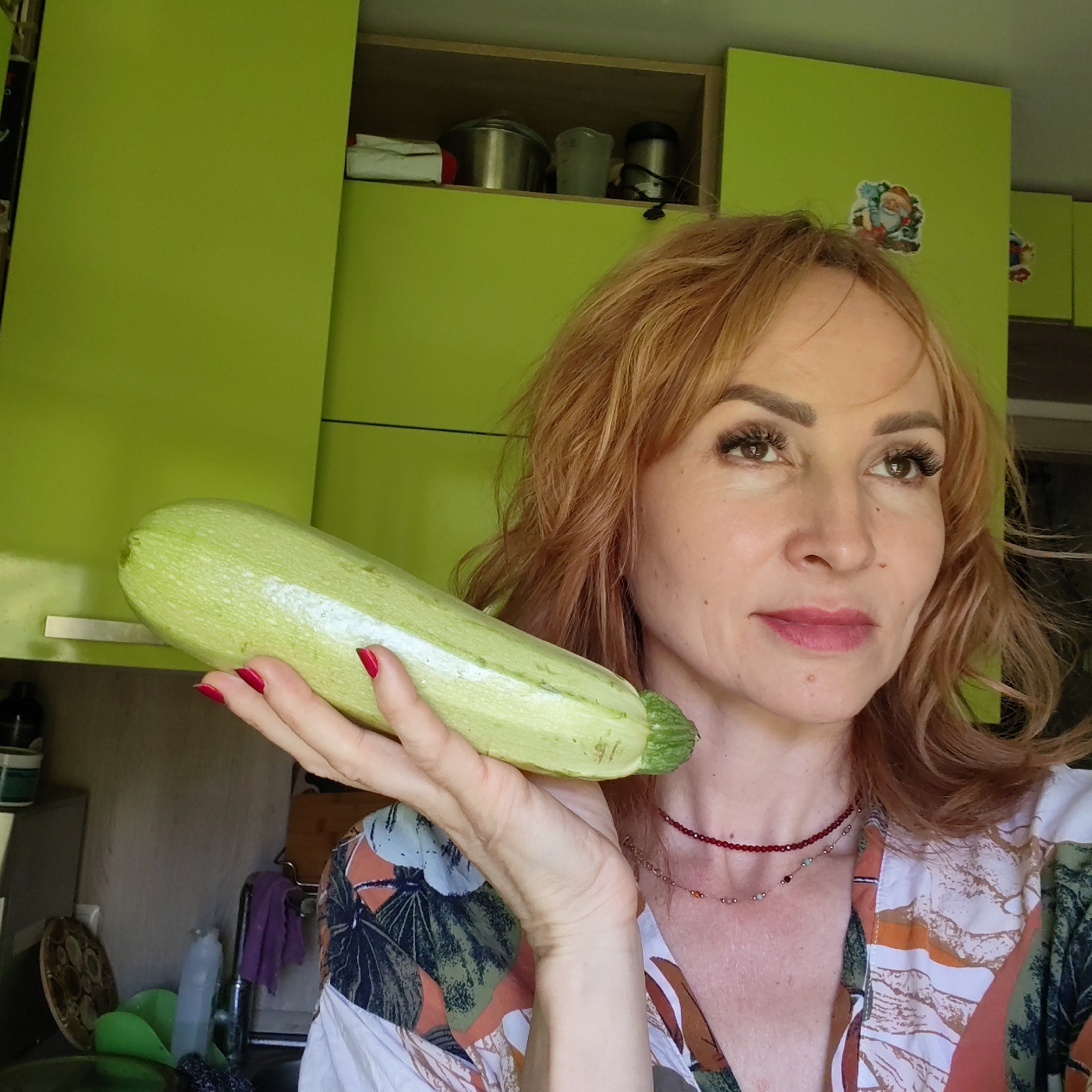 A woman with shoulder-length reddish-blonde wavy hair holds a light green zucchini in her outstretched right hand. She has fair skin, visible eyelashes, and wears a pink lipstick. Her fingernails are painted red. She wears a patterned shirt and two necklaces. The background includes light green kitchen cabinets, some with stickers on them, and kitchen utensils inside an open cabinet. The location is presumed to be a kitchen in Chaikovsky, Russia, although there is no conclusive evidence in the image to confirm the city.