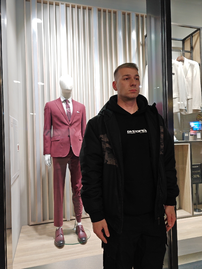 A scene within a retail clothing store in Shumen, Bulgaria. In the immediate foreground, a light-skinned male with short, light brown hair and a neutral expression stands facing slightly right. He is wearing a black hooded sweatshirt with "DYSTOPIA" printed in white on the chest, a black unzipped jacket with dark camouflage shoulder accents, and black trousers. His left hand is visible at his side. Behind him and to his left, inside a display window, a white male mannequin is dressed in a fitted maroon suit, white shirt, maroon tie with a silver tie clip, and dark red oxford shoes. The display area behind the mannequin features vertical light-colored panels. To the right, further inside the store and visible through glass, a shelving unit displays light-colored suits on hangers, with additional clothing items and a sign indicating "-30%" off. The flooring throughout the visible retail space is light-colored.