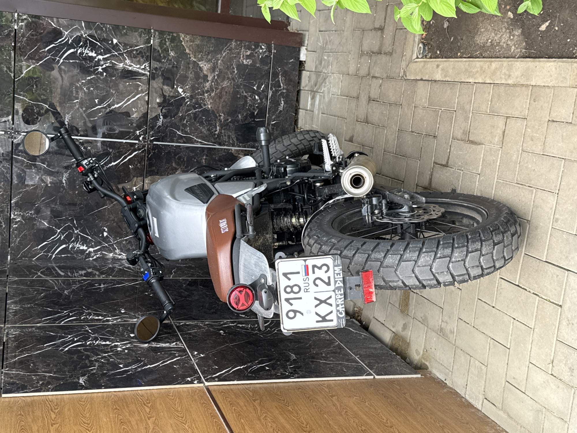 The image depicts a SYM motorcycle parked on a tiled surface. The motorcycle is gray and black with a brown seat, and has a license plate with the number "9181 KX 23" which indicates registration in the city of Krasnodar, Russia. The motorcycle is surrounded by tiled surfaces, including a dark marble pattern behind it and light gray brick-pattern tiles to its right. In the upper right corner, there is some green foliage. In the foreground, there is an area with wood-like tiles.