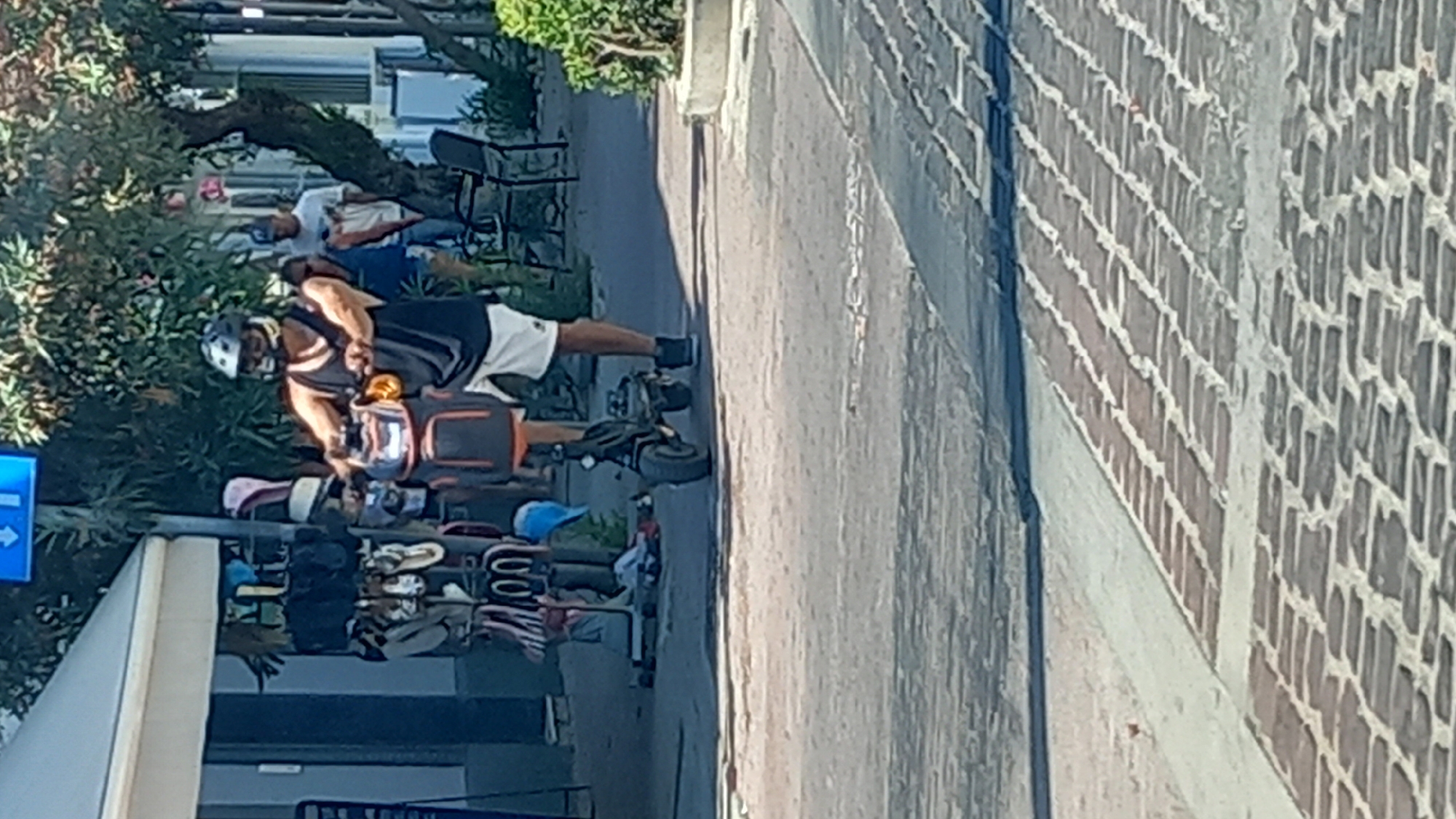 A man wearing a helmet and a black tank top rides a black electric scooter with an orange and black backpack attached to the handlebars. He is on a paved walkway lined with brickwork. To his left, there is a stand with various hats and sandals displayed. Behind him, trees and other pedestrians are visible. The scene appears to be in the city of Kos, Greece, likely in a tourist area with shops and vendors.