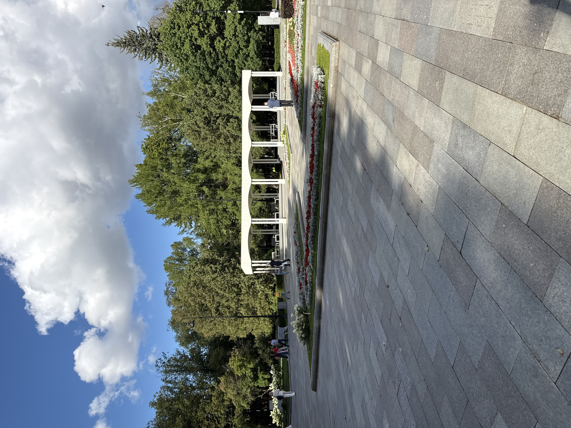The image depicts a sunny day in a park located in Khimki, Russia. The foreground is a wide expanse of grey stone paving, with a subtle pattern of shadows cast across it. In the midground, a white, open-air pavilion with multiple arches and supporting columns stands prominently. Surrounding the pavilion are flowerbeds filled with red and white flowers, bordered by green grass. Several people are visible in the scene: a woman in white walks away from the camera on the left, a group of people stand and walk near a bench in the distance, and a man in a blue shirt stands facing the pavilion. Behind the pavilion and flowerbeds, a dense line of lush green trees forms the backdrop, with a few taller coniferous trees interspersed. The sky above is a vibrant blue, dotted with fluffy white clouds. The overall impression is one of a well-maintained public space enjoying pleasant weather.