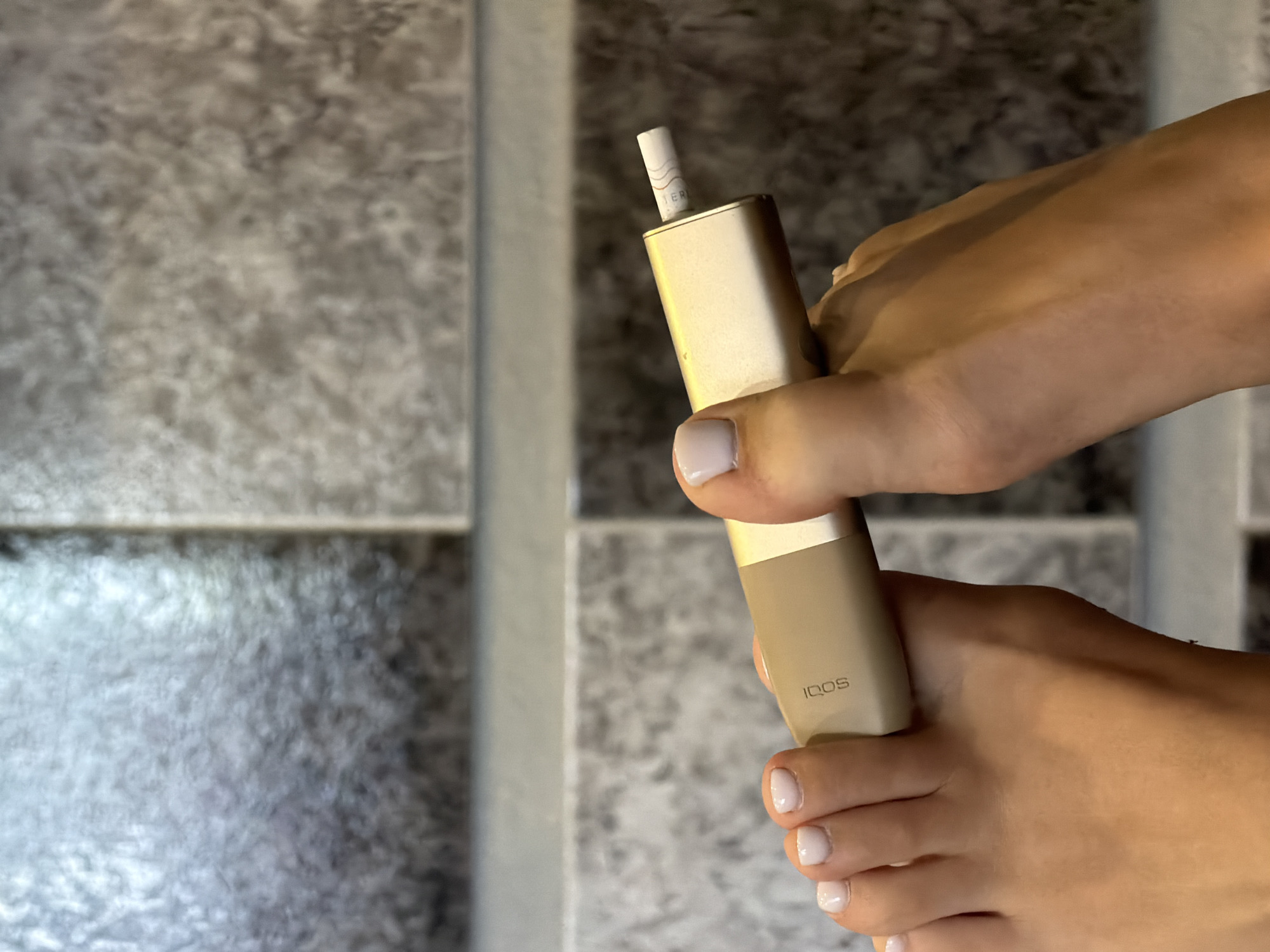 In Novi Iskar, Bulgaria, a close-up shot focuses on a pair of human feet, one on the left and one on the right, holding a golden-colored electronic smoking device. The device is positioned horizontally between the toes. The background consists of textured gray tiles, arranged in a grid pattern.