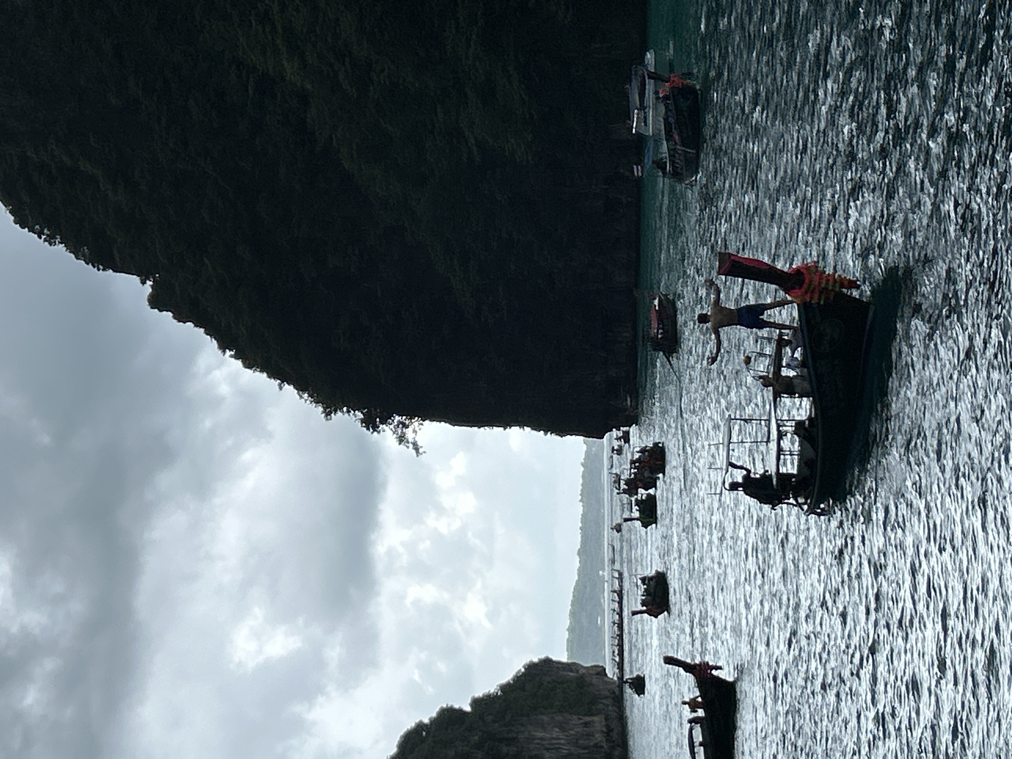 The image depicts a panoramic view of a bay in Haad Yao, Thailand, under a cloudy sky. Several long-tail boats are scattered across the turquoise water, with two large, tree-covered cliffs framing the scene. In the foreground, a long-tail boat carries three people, one standing with arms outstretched and another sitting. Other boats are visible in the middle ground and background, some with passengers. The water exhibits moderate ripples, indicating gentle wave activity. The overall mood is serene yet dramatic, owing to the overcast sky and the imposing cliffs.