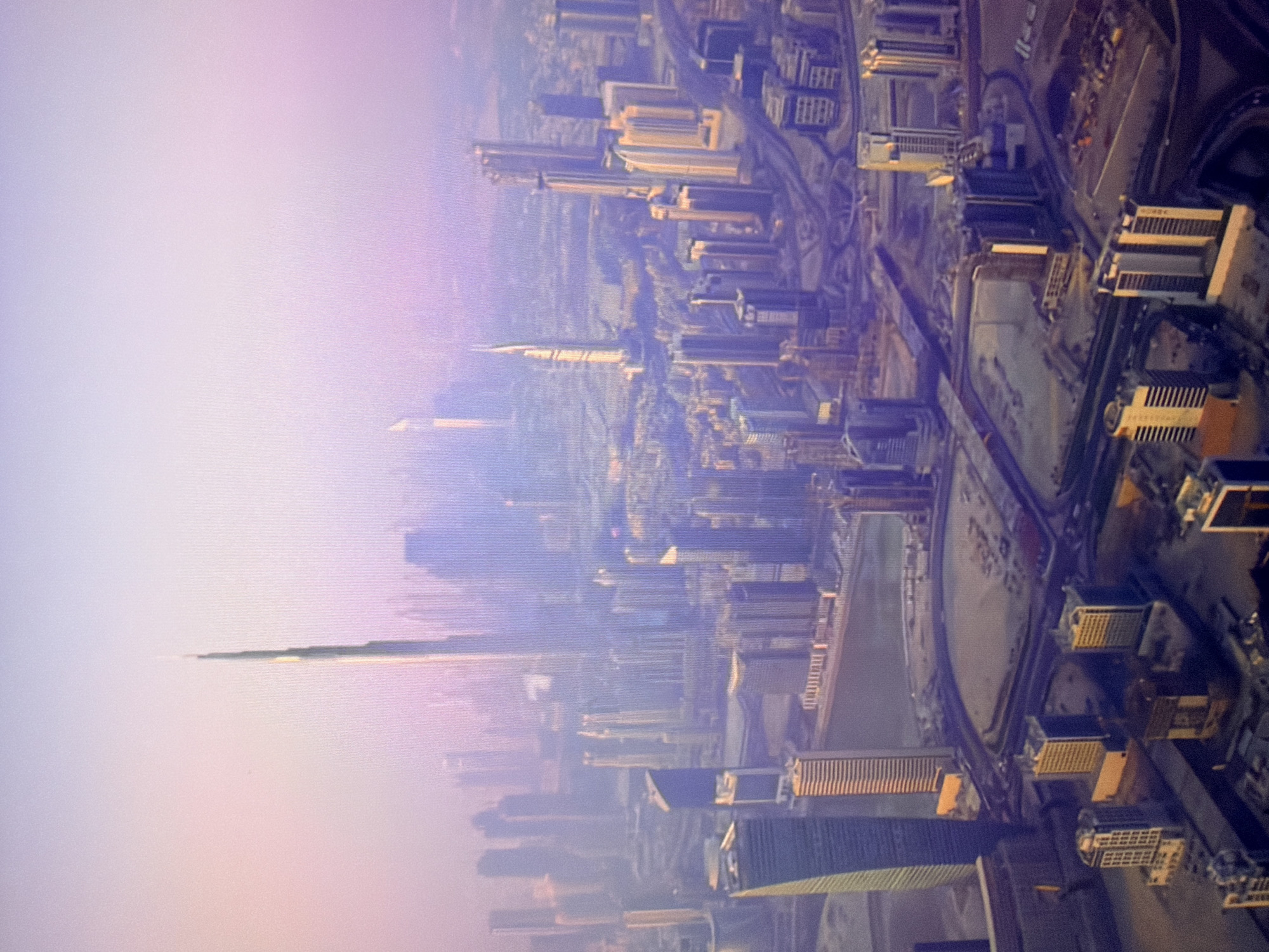 The image is an aerial view of a modern city skyline, dominated by numerous skyscrapers of varying heights and architectural styles. The tallest and most prominent structure, a needle-like skyscraper, is positioned in the upper central part of the frame, catching the light. Other buildings form dense clusters, with some appearing to be under construction or recently completed, suggested by the exposed earth and construction sites visible in the foreground. Roads and elevated highways snake through the urban landscape, connecting different parts of the city. The atmosphere appears hazy, with a soft, pastel-colored sky, possibly indicating dawn or dusk, or atmospheric conditions like dust or fog. The overall impression is one of a sprawling, contemporary metropolis.
This image does not depict the city of Kyiv, Ukraine. The architecture, particularly the prominent skyscraper in the center, is characteristic of cities like Dubai, United Arab Emirates.
