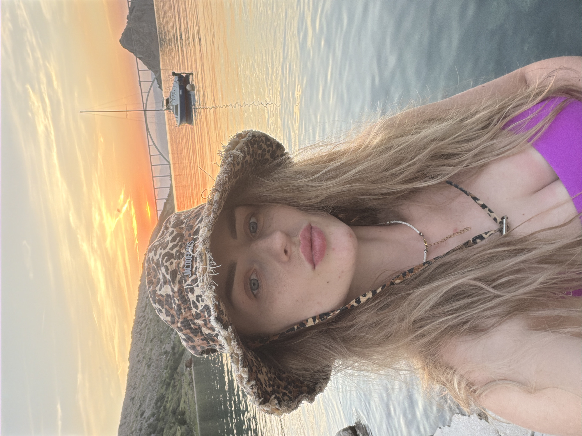 A woman is taking a selfie in front of a body of water with a sunset in the background. She is wearing a leopard print bucket hat and a pink bikini top. Her hair is long and blonde. In the background, there is a sailboat and a bridge. The sun is setting, casting an orange glow over the scene. The water is calm and reflects the colors of the sunset. The location is unknown.