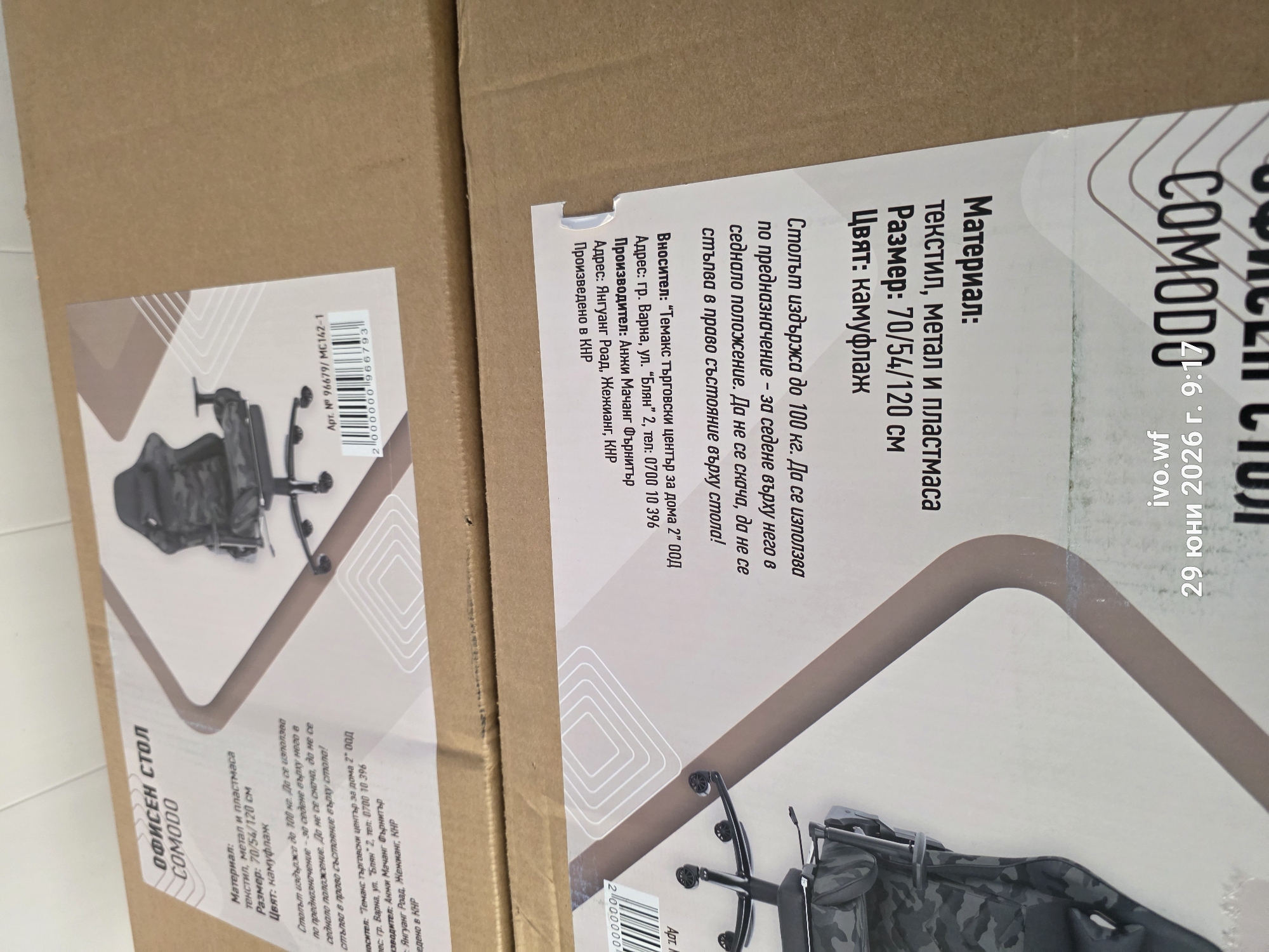 This image shows a cardboard box with text and graphics printed on it. The text is in Bulgarian and describes an office chair named "COMODO". The graphics include an illustration of the chair and a barcode. The box appears to be part of a product shipment or inventory. The bottom right corner of the image contains the text "ivo.wf 29 юни 2026 г. 9:17", which is likely a timestamp or identifier related to the photo. The box is placed on top of another cardboard box, and the overall environment suggests a storage or shipping facility. There are no people or specific locations in the city of Chal, Bulgaria, depicted in this image.