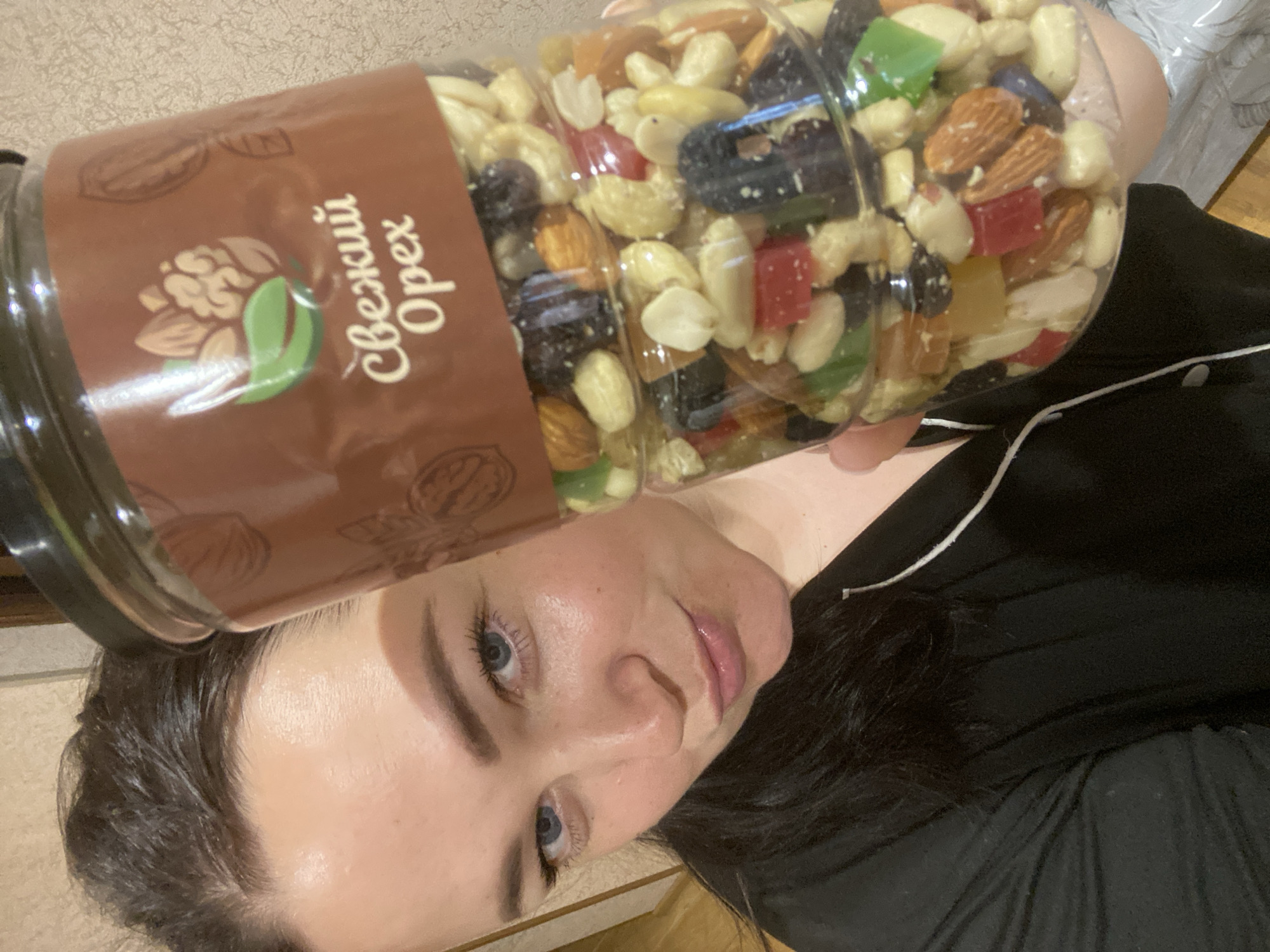 The image shows a woman holding a plastic jar of mixed nuts and dried fruits above her head. The jar has a brown label with a walnut and almond illustration and the text "Свежий Орех" in Cyrillic, translating to "Fresh Nut" in English. The mix visible inside the jar includes cashews, almonds, peanuts, and pieces of dried fruits with colors such as red, green, yellow, and blue. The woman is fair-skinned with blue eyes and dark hair. She wears a black top with a white trim along the neckline. The background is a blurred beige surface, possibly a wall or wallpaper. There is no evidence to suggest that the photograph was taken in Sochi, Russia.