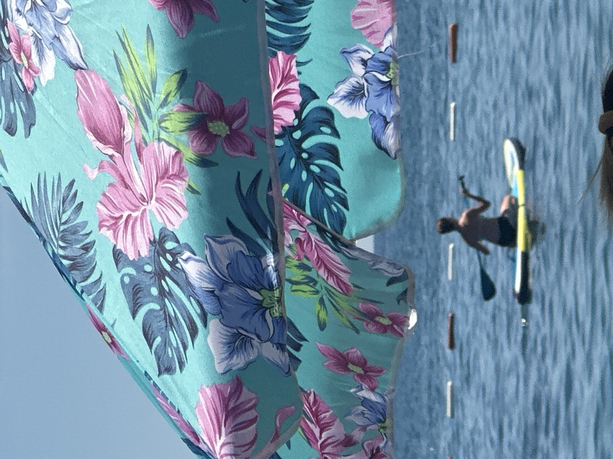 The image captures a sunny day scene at a body of water, likely a beach or bay, in Boreti, Montenegro. In the foreground, a large, turquoise beach umbrella with a vibrant floral pattern featuring pink and blue flowers and dark green leaves dominates the upper portion of the frame. The umbrella partially obscures the view of the water.
In the middle ground, a person is seated on a yellow and blue paddleboard in the blue water. The individual is facing away from the camera and is holding a paddle, appearing to be in the process of paddling. Several buoys, some white and one brown, are floating in the water, marking an area.
The background consists of the clear blue sky and the rippling surface of the water. The overall atmosphere is one of leisure and outdoor activity.