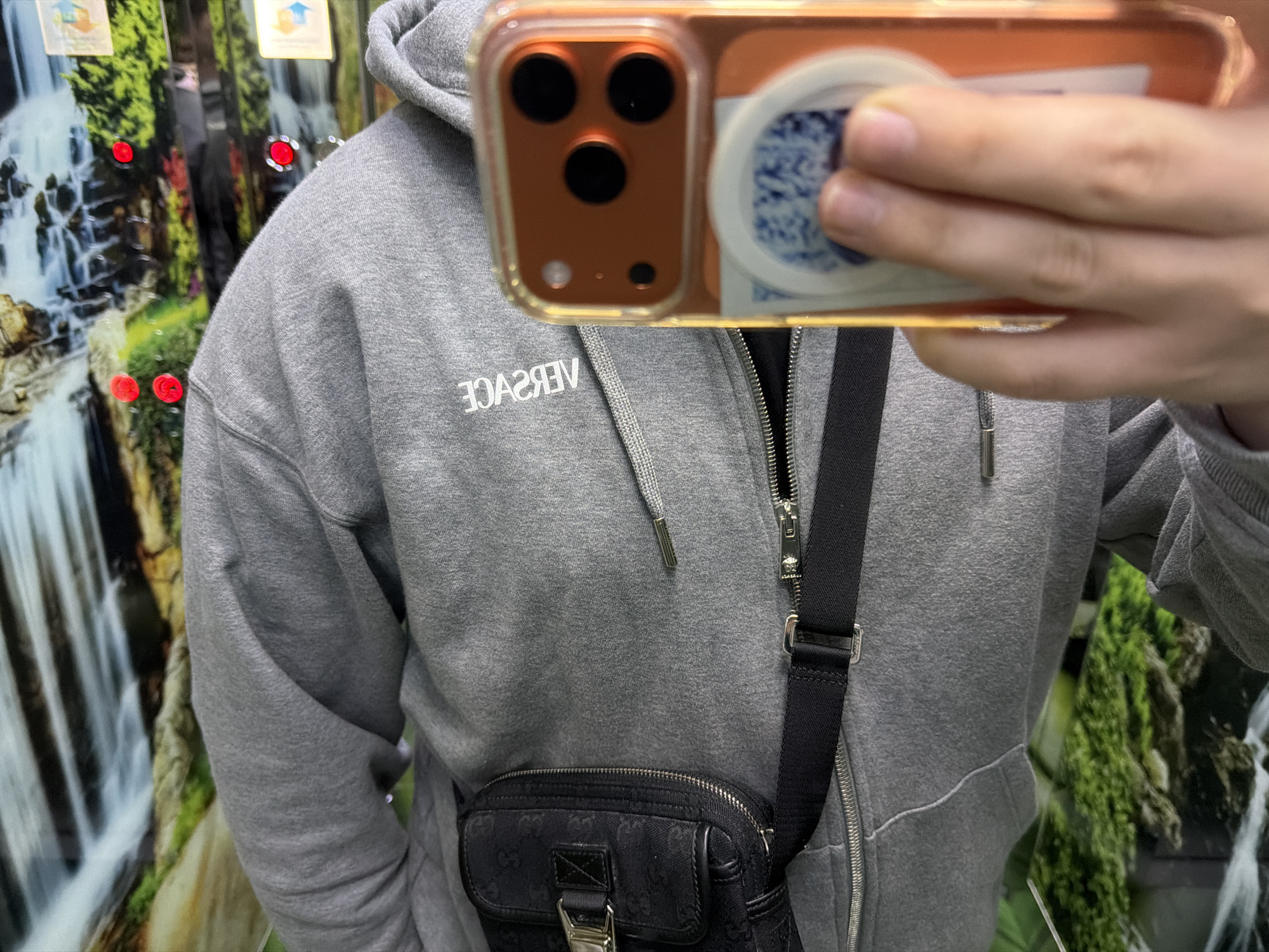A person wearing a gray Versace hoodie is taking a mirror selfie with an orange iPhone. The background appears to be a shop display or possibly an elevator with a nature scene backdrop depicting a waterfall and greenery. The person is also wearing a black Gucci messenger bag slung across their body. The location in the city of Budva, Montenegro is not clearly discernible from the image.