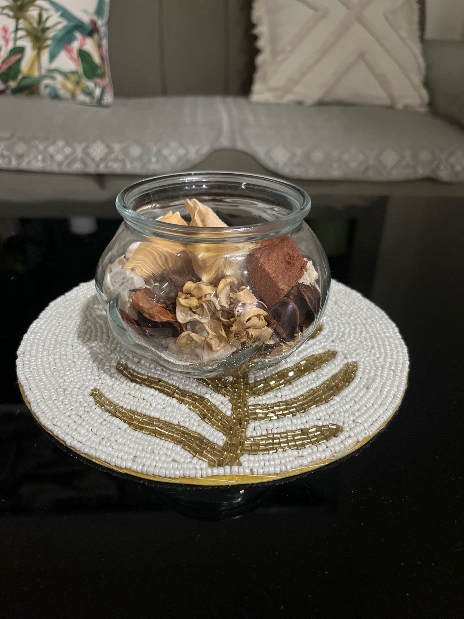 The image shows a close-up of a glass bowl filled with potpourri, placed on a beaded placemat with a gold leaf design. The placemat rests on a dark, reflective surface, likely a coffee table. In the background, a sofa with decorative cushions is visible, suggesting an indoor setting. The potpourri consists of dried natural elements like flower petals and wood pieces in shades of beige, brown, and red. The beaded placemat is predominantly white with gold accents forming a leaf pattern. There are no people or animals present in the image. The lighting appears to be ambient, creating soft shadows. The location in Firozpur, India, is not discernible from the visual information provided in the image.