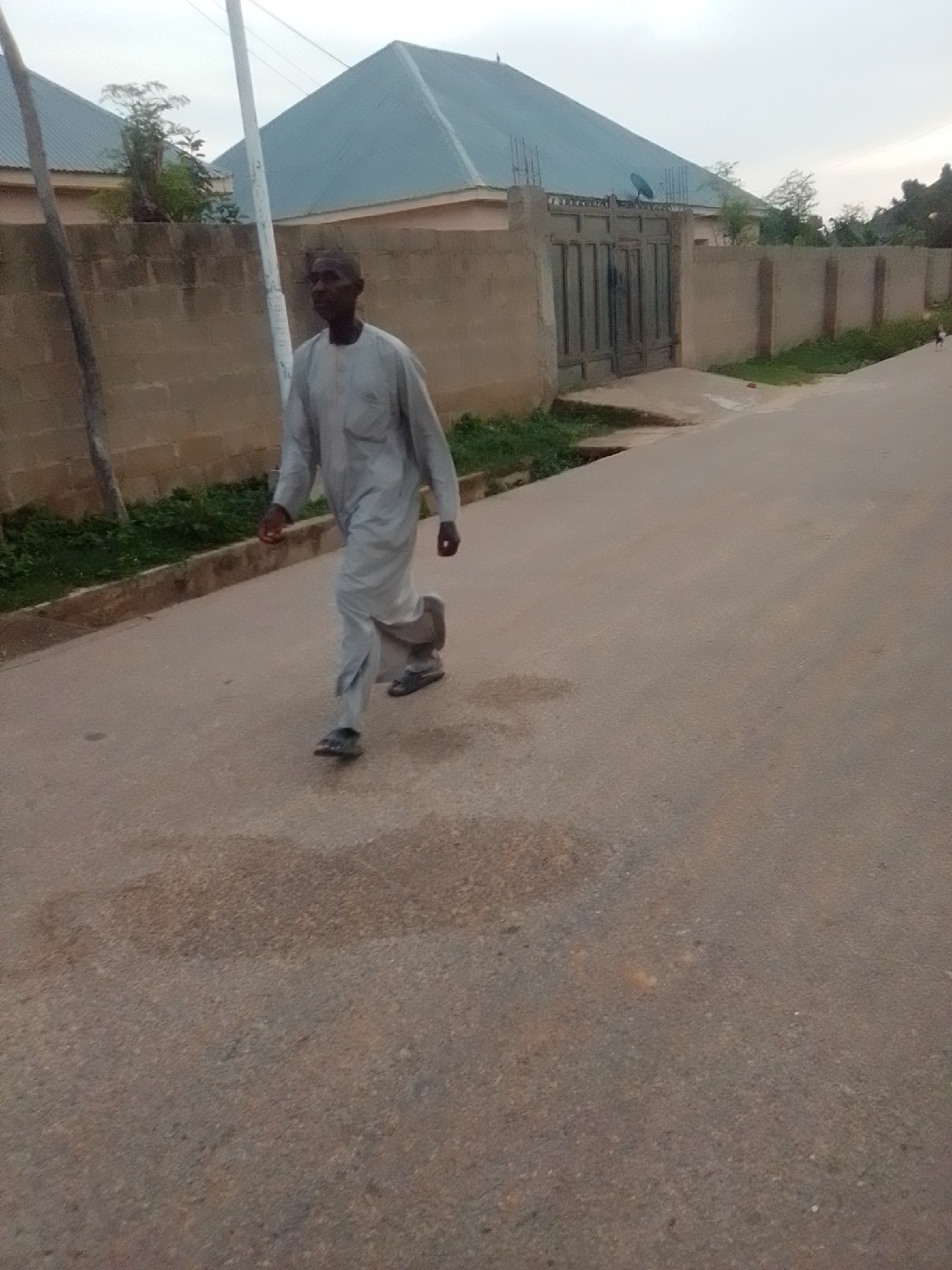 A man in a light grey traditional outfit and dark sandals walks on an asphalt road, his body angled towards the viewer's left. He is in motion, with his left leg forward and his right leg trailing. Behind him, a tan brick wall topped with a lighter colored coping encloses a property with buildings that have blue corrugated metal roofs. A dark metal gate is visible within the wall. Green foliage lines the base of the wall. A utility pole stands to the left of the man. The sky is overcast and light. The overall scene appears to be a street in a residential area.