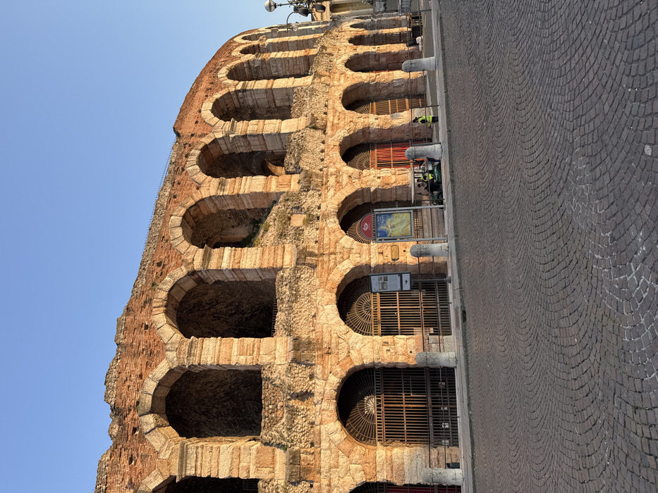 The image displays the exterior of the Arena di Verona, situated in Piazza Bra, Verona, Italy. The ancient Roman amphitheater, constructed from light-colored stone and reddish-orange brick, occupies the upper two-thirds of the frame, featuring multiple tiers of arched openings. The lower level arches are predominantly secured with dark metal grates, some showing glimpses of red elements within. Two public information kiosks, one rectangular and one with an arched top displaying a colorful map, are positioned in front of the lower facade. A low, light-colored stone barrier with horizontal metal rails runs along the base of the arena. In the foreground, a wide expanse of dark grey cobblestone pavement is visible, arranged in a radial pattern. On the right side, a dark green utility vehicle, resembling an electric golf cart, is parked behind the barrier. Two individuals are present: one stands next to the utility vehicle, facing left and wearing a bright yellow high-visibility vest, and at least one other person, also in a high-visibility vest, is seated inside the vehicle. A multi-headed dark lamppost is partially visible on the far right. The scene is illuminated by natural sunlight under a clear, bright blue sky.