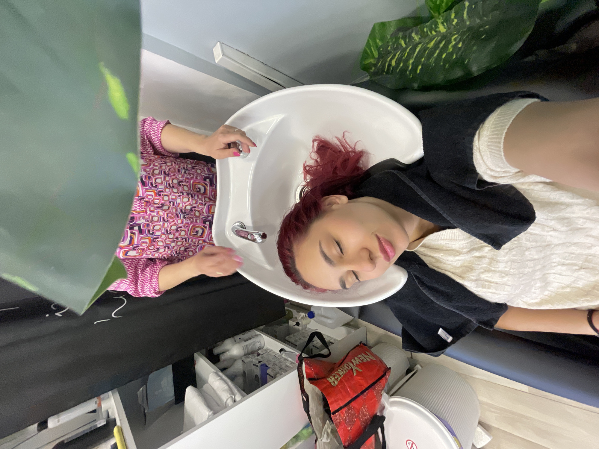 In an indoor scene, likely a hair salon in Pleven, Bulgaria, a woman with vibrant red hair is reclined at a shampoo sink, appearing relaxed with her eyes closed, as another person's hands (wearing a pink patterned top and pink nail polish) wash her hair. Her red hair is gathered in the white ceramic sink, which has a silver faucet. She is wearing a cream-colored, textured sweater under a black cape or towel draped over her shoulders for protection.
The perspective is somewhat angled and elevated, capturing the woman's face and the shampoo station. In the background, behind the sink, a large green leafy plant is visible. Below the sink, various salon supplies are neatly stored in white drawers, including bottles, boxes, and white towels. A red bag with the text "NEW YORKER" in white is also visible near the drawers, along with a white ribbed bin or bucket. The floor appears to be light-colored wood or laminate. The overall ambiance suggests a typical salon setting during a hair treatment.