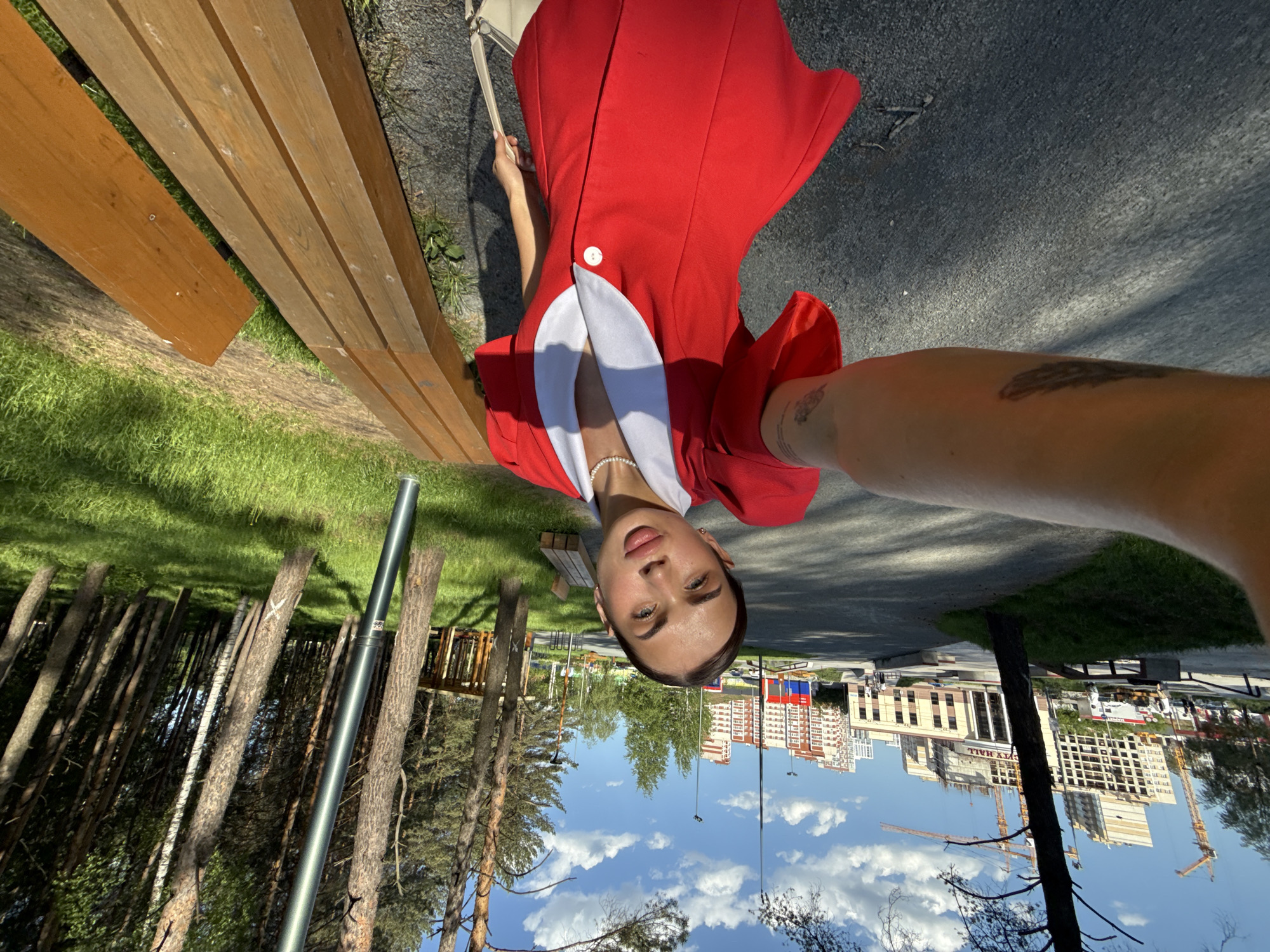 A woman wearing a red dress with white details is centered in the frame, reclining against a wooden bench. Her face is prominent, facing the camera. The photograph appears to be a selfie.
The background includes a park setting with trees and grass. In the distance, buildings and construction cranes are visible. The architecture and cityscape suggest the photo was taken in an urban area. Based on the presence of Russian flags and architecture, the location is likely Perm, Russia.