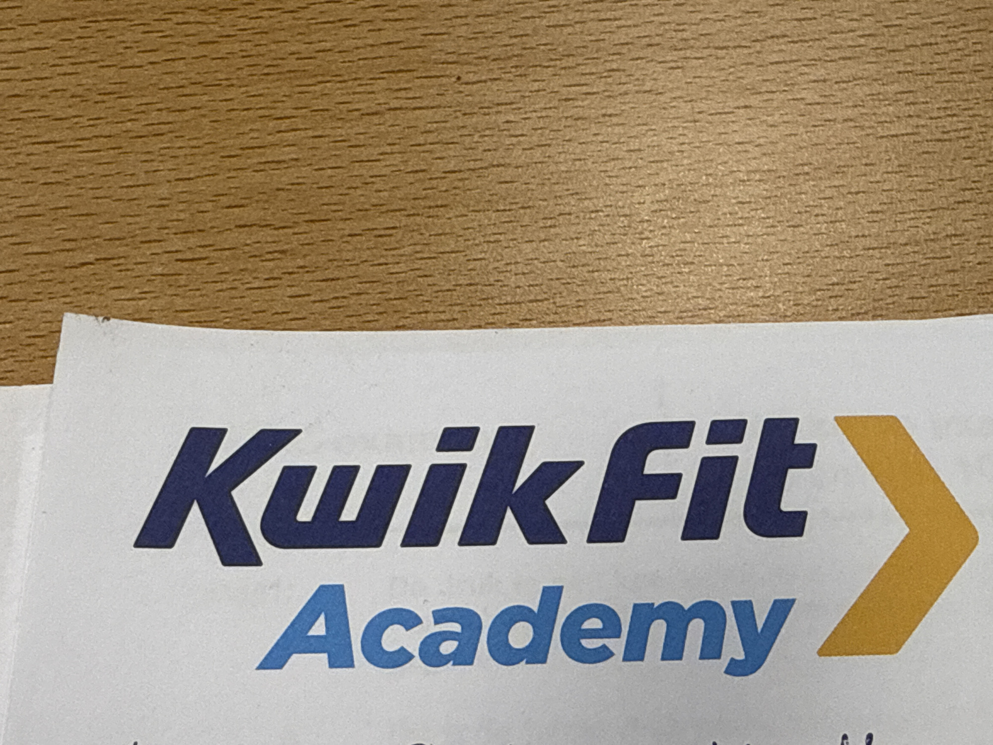 The image shows a portion of a printed document with the words "Kwik Fit Academy" written vertically in bold, dark blue capital letters. Below the text is a large, downward-pointing yellow chevron. The document is placed on a light-colored surface and is angled to the right, with a textured, light brown wooden surface visible to the right of the document. No people or interactions are visible. The location of Harderwijk, Netherlands, is not depicted in the image.